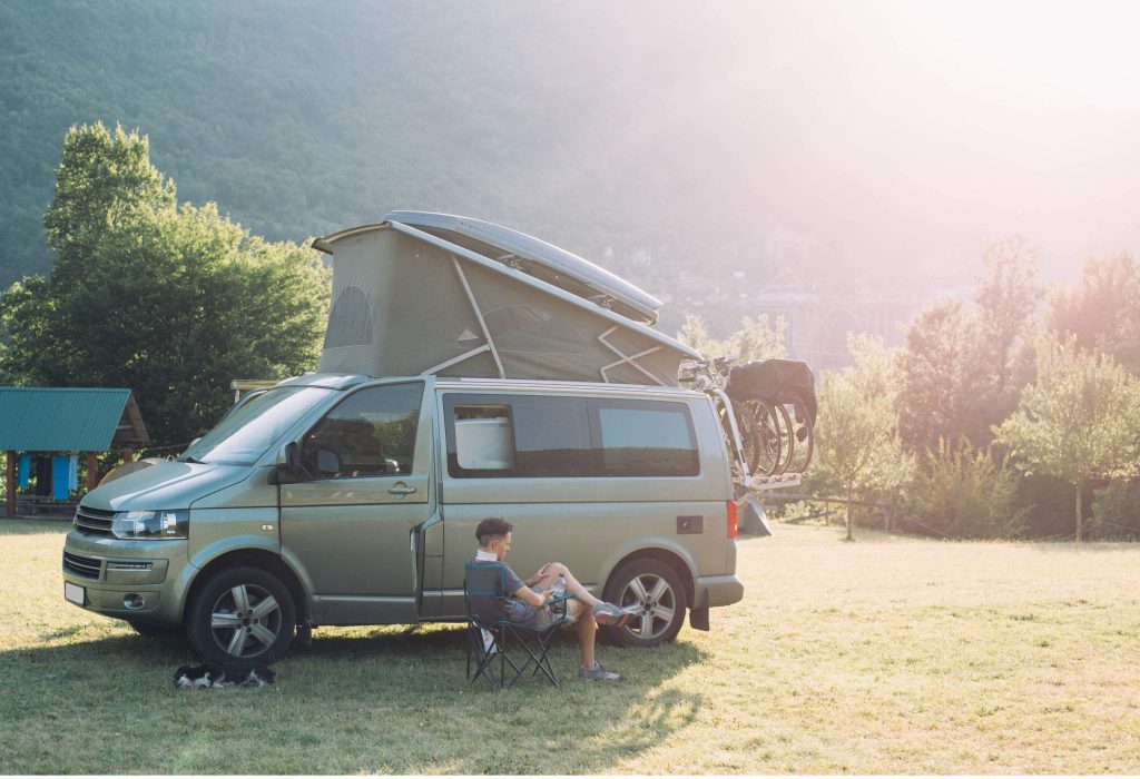 A person sits on a folding chair next to a sleeping dog and a grey camper van equipped with a bike rack and a rooftop tent.