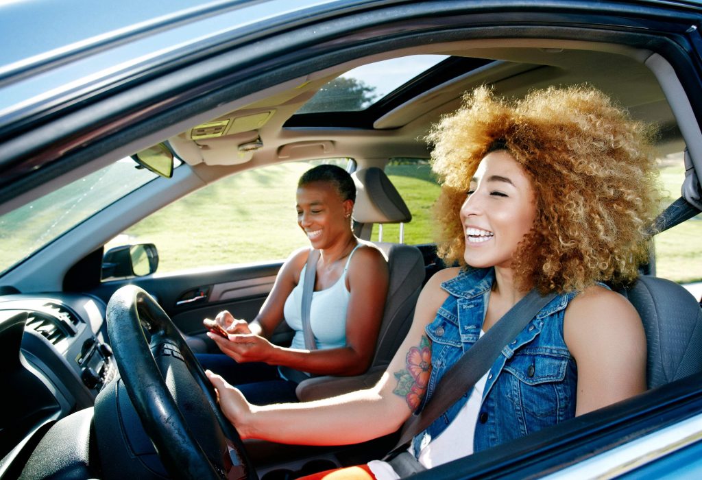 A woman with kinky blonde hair driving a car with a friend sitting on the passenger seat.