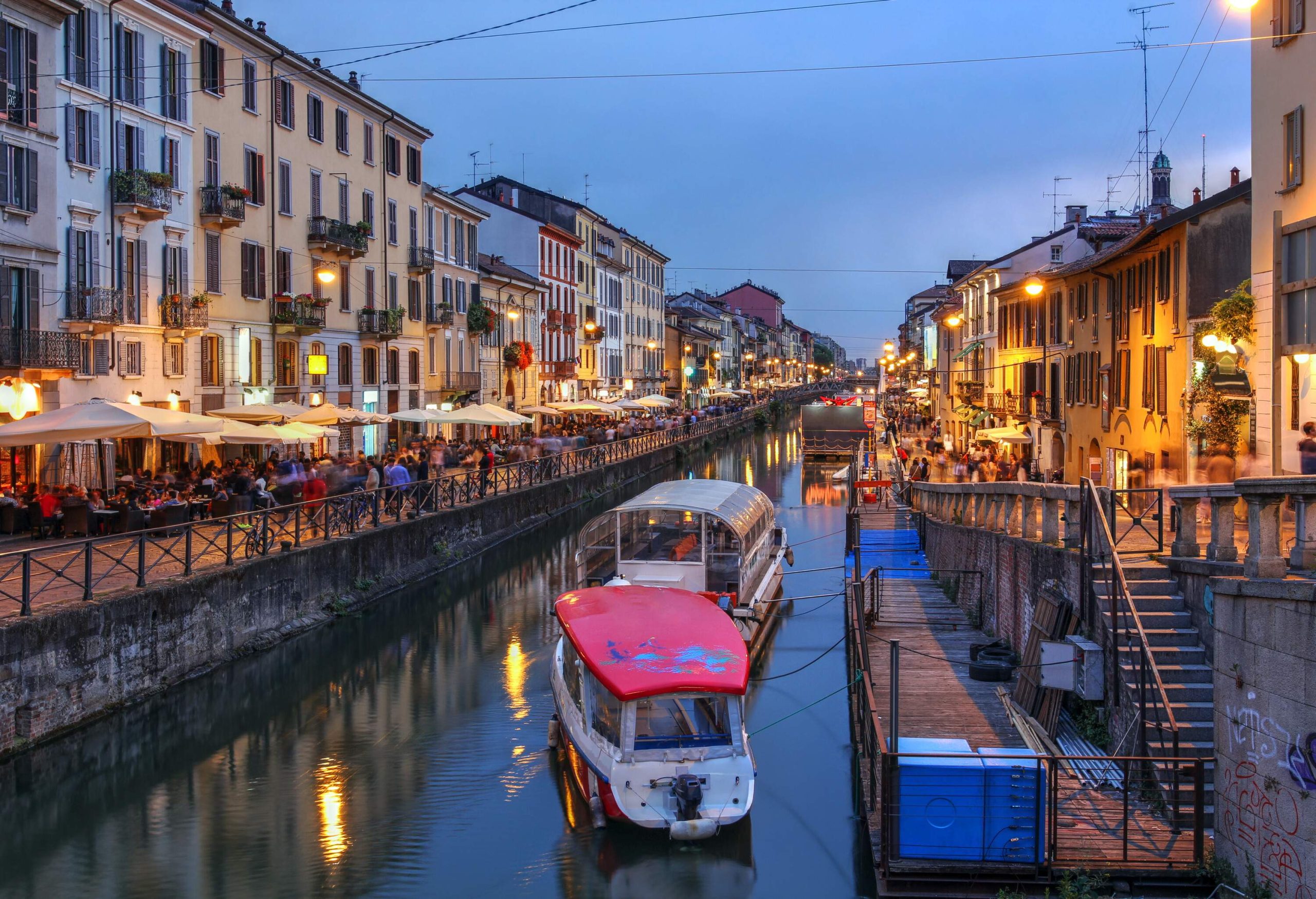 A canal pass through in the middle of illuminated, crowded promenades lined with buildings.