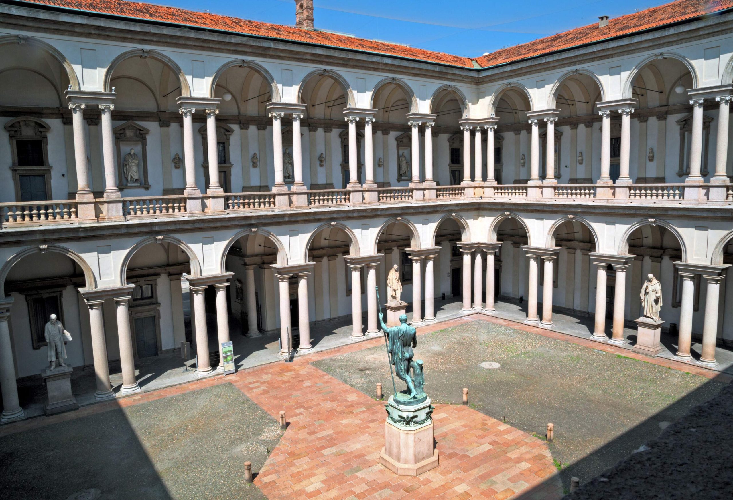 A courtyard with a statue in the centre and along the columns of a two-storey arcade building.