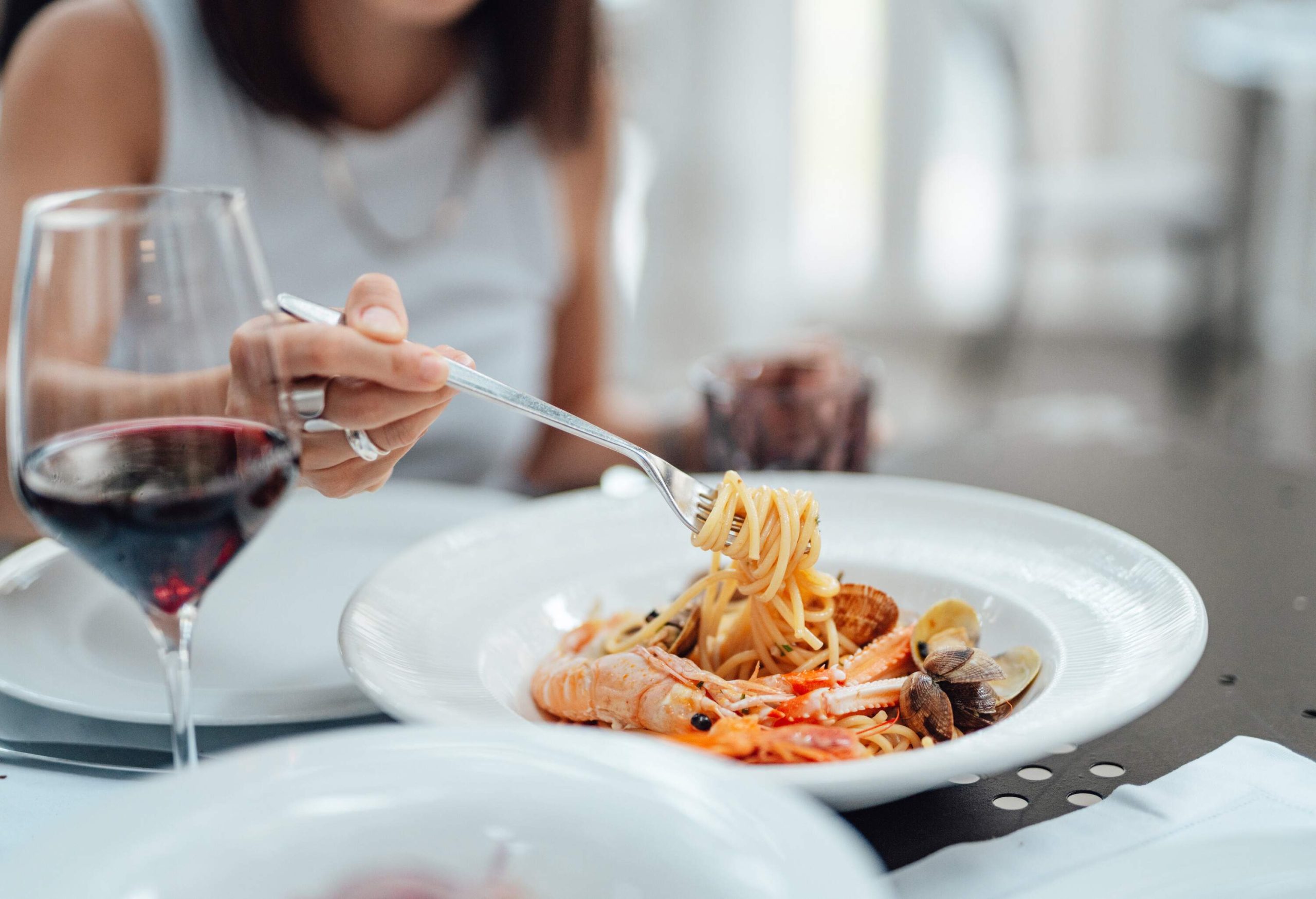 A woman is picking from a plate of seafood pasta next to a glass of red wine.