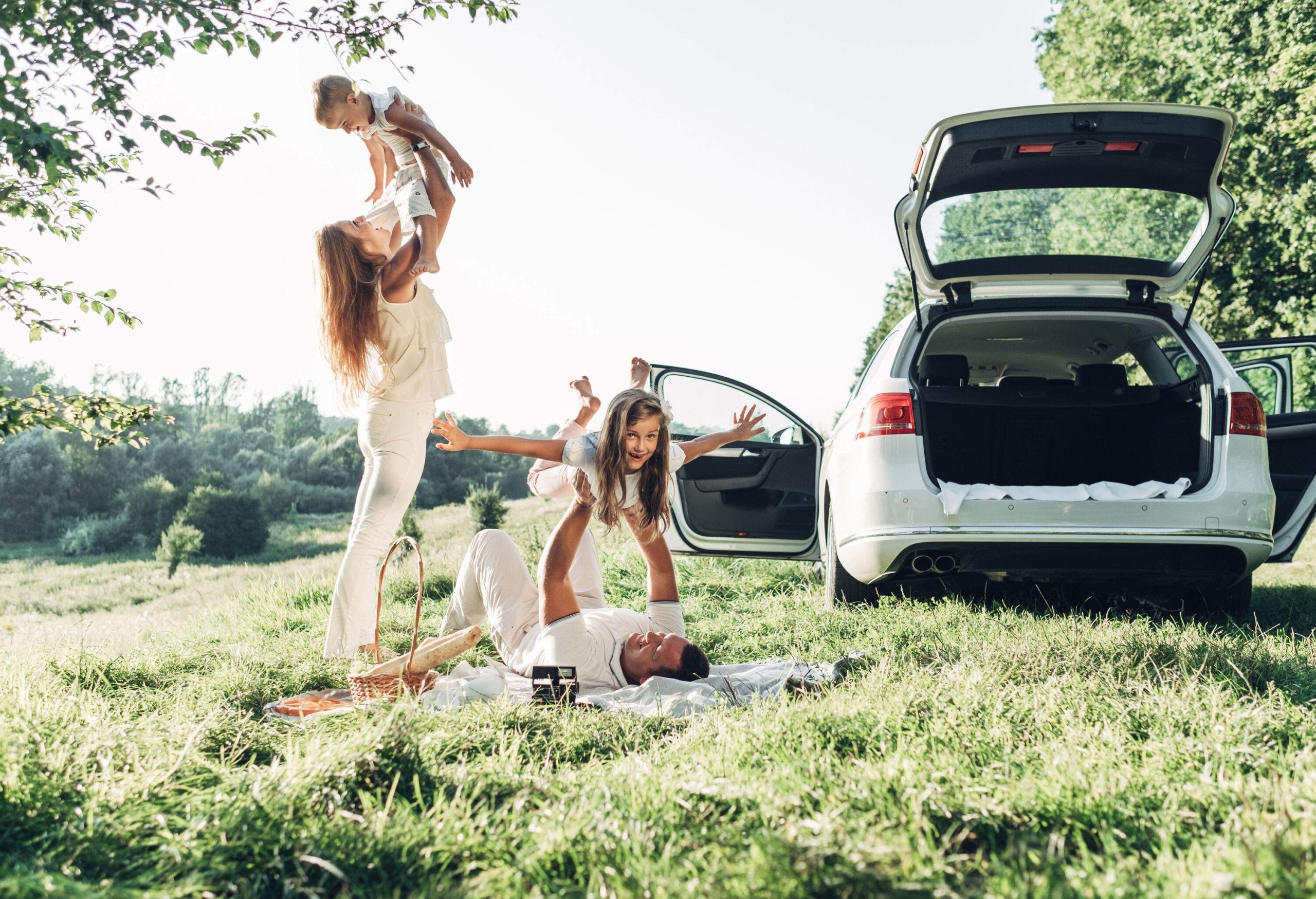 A woman stands while hoisting a boy into the air next to a man lying on a blanket lifting a girl beside a white car.