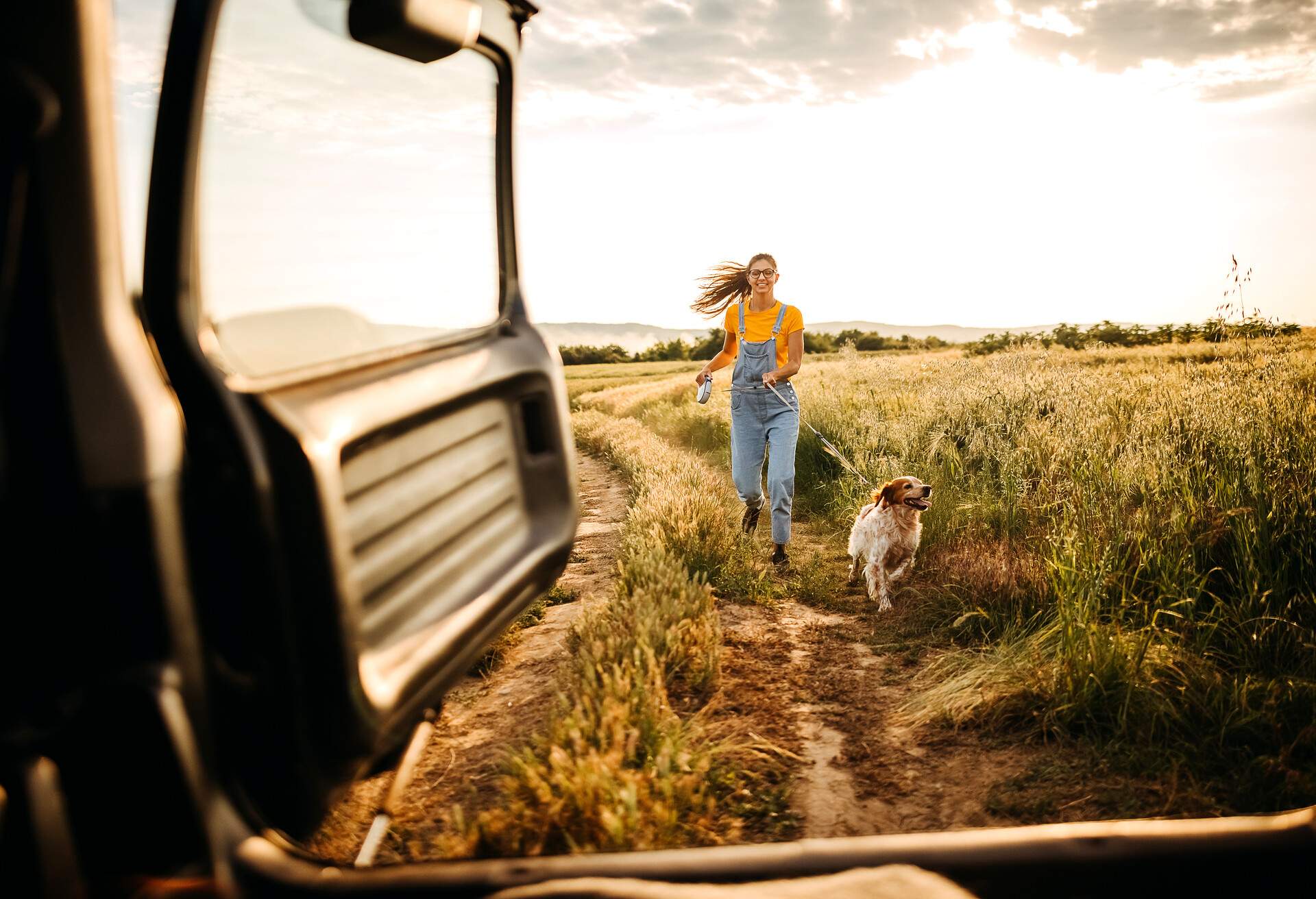 A woman with a dog hold by a leash, in a field, joining the car to continue the road trip