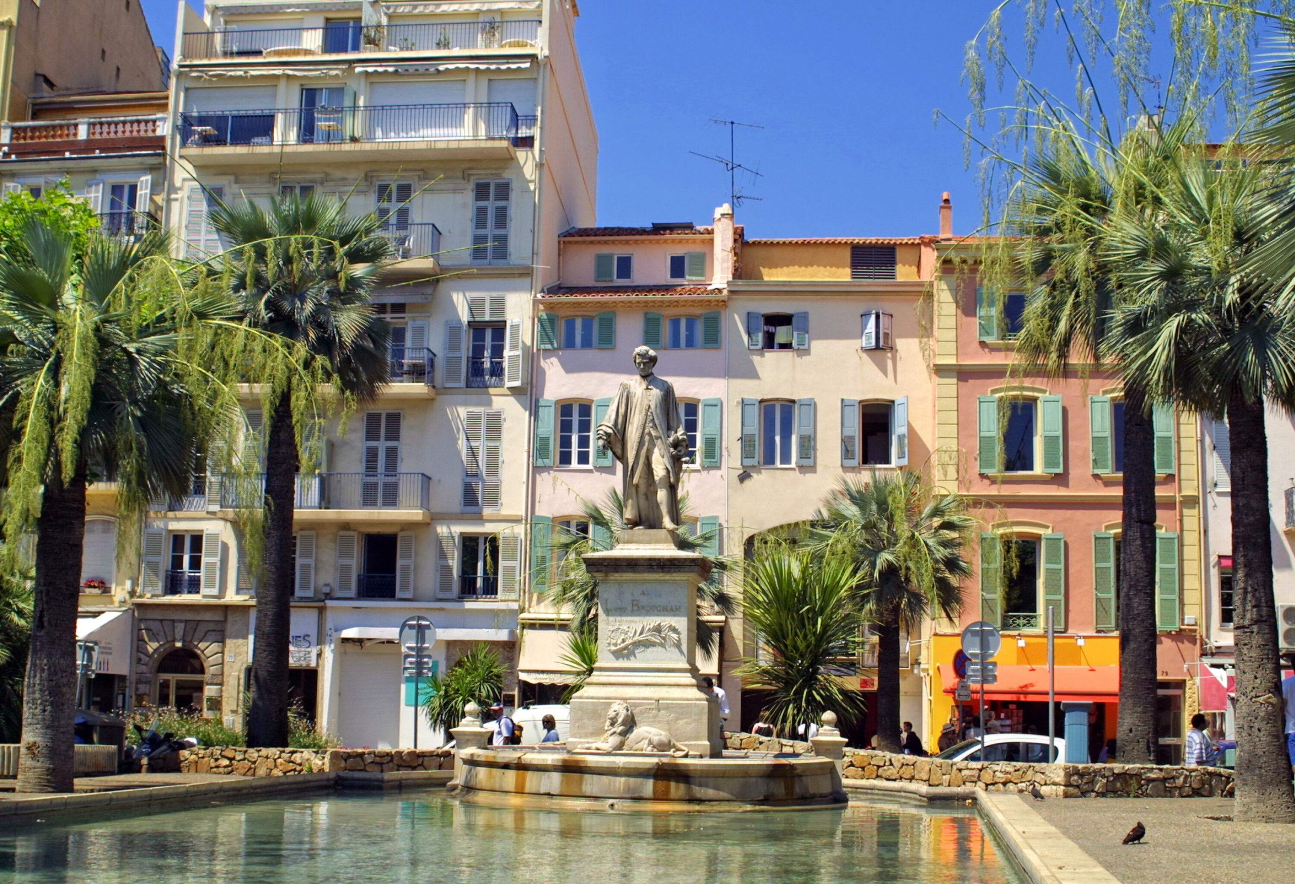 A statue above a lion over a pond surrounded by palm trees alongside a line of colourful buildings.