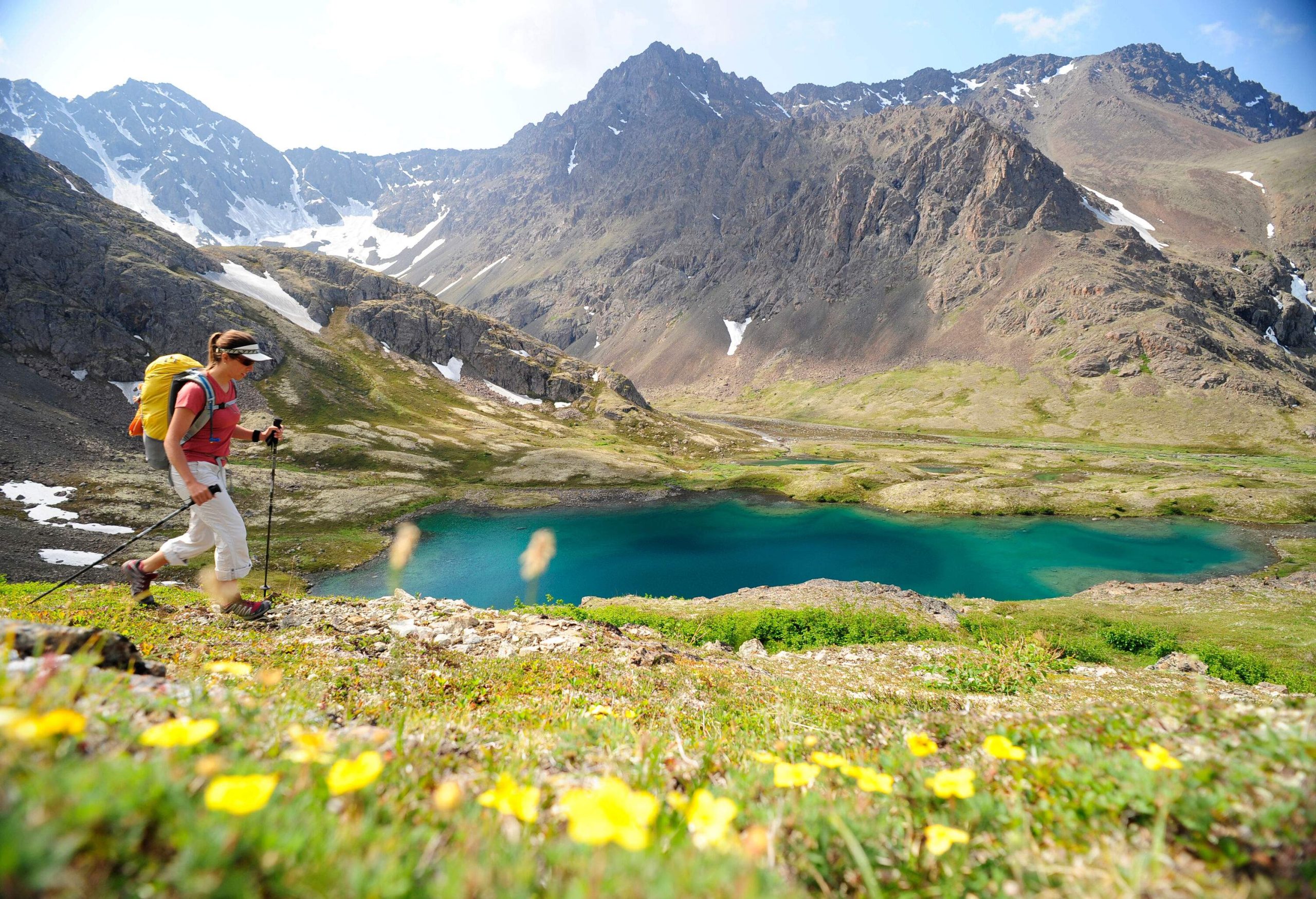 A female hiker walking along the coast of a blue lake beneath the tall mountains.