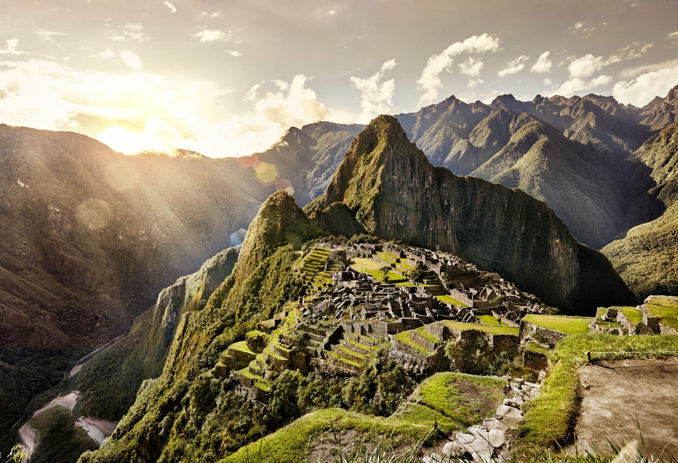 The brilliant sun bursting through the open sky, shedding light on the mountains surrounding the Machu Picchu.