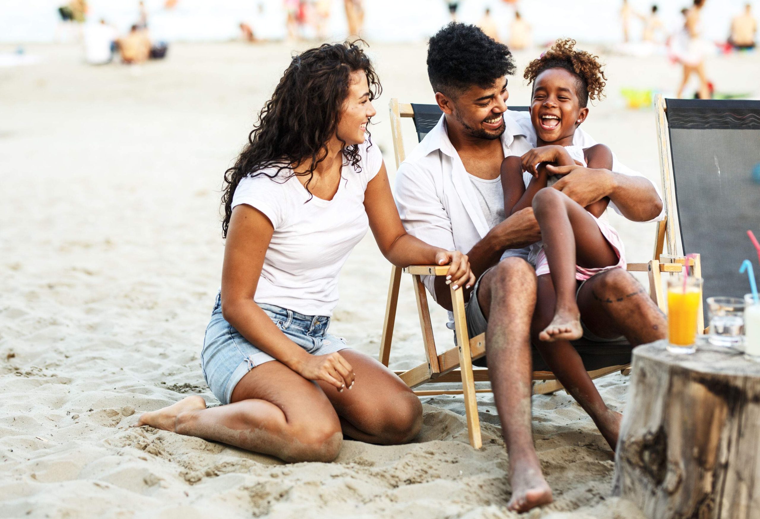 A man sits in a chair with a laughing child on his lap, next to a woman sitting on the sand.