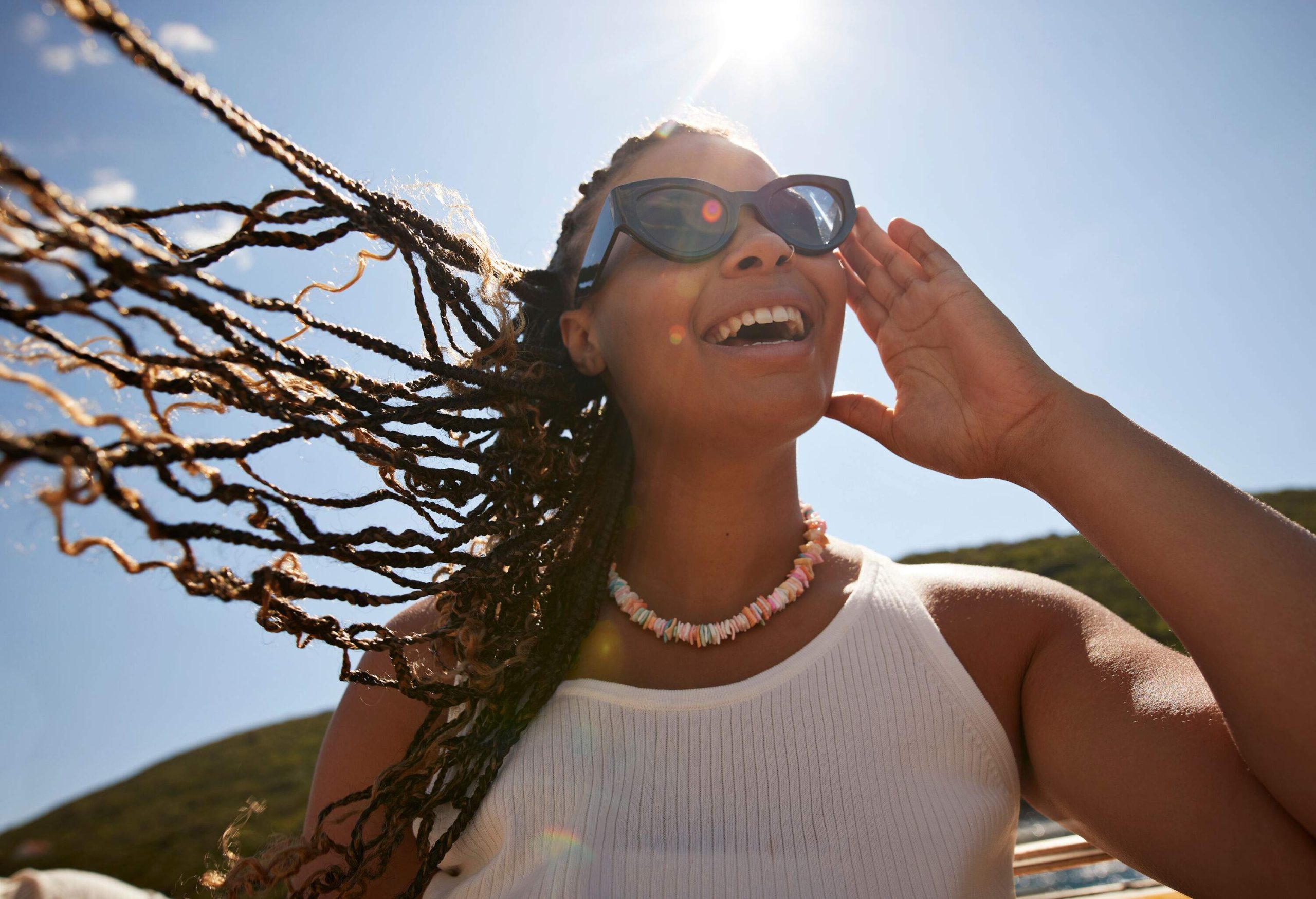 Happy young woman with tousled braided hair adjusting sunglasses during summer vacation on sunny day