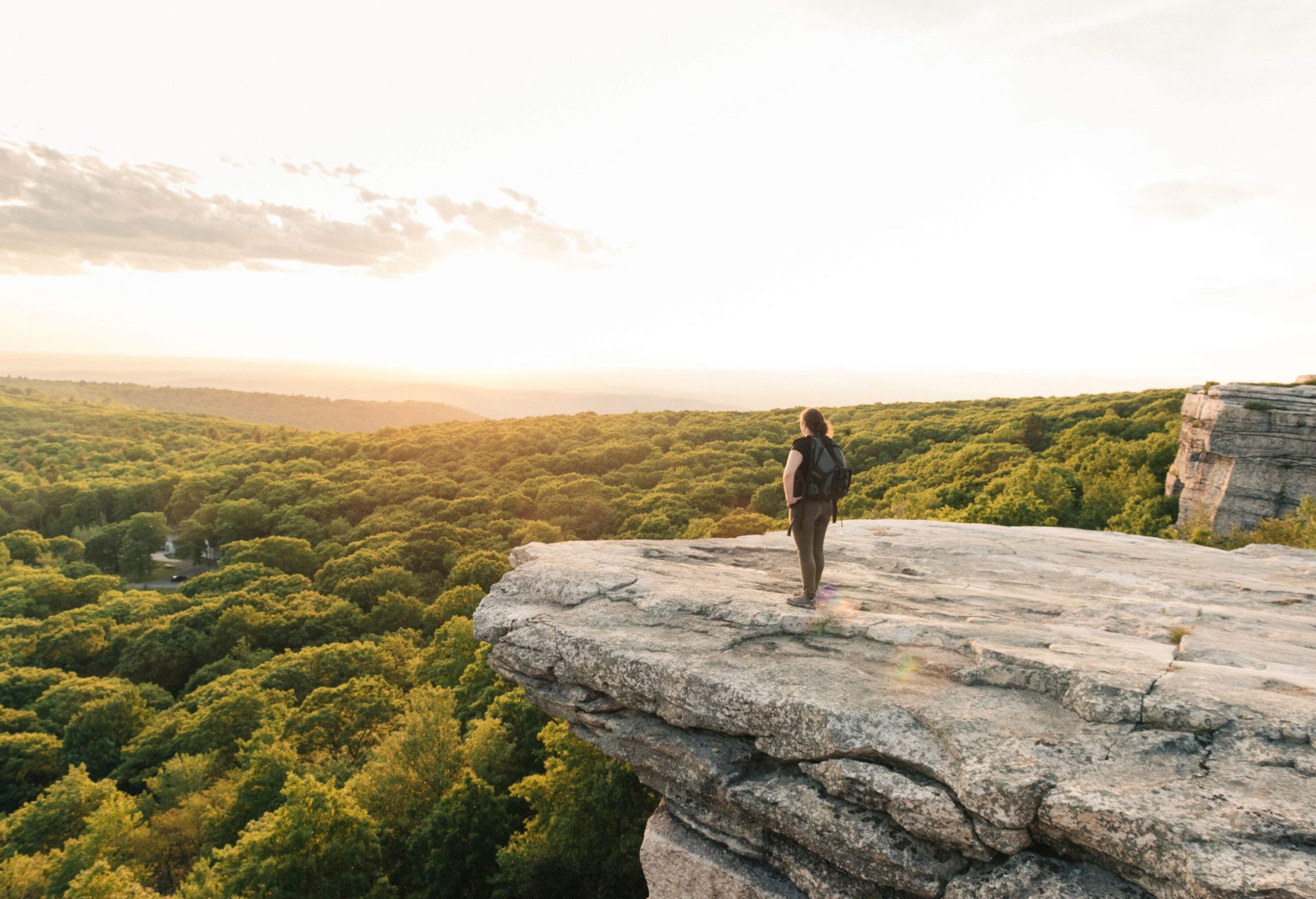 A person standing on a cliff's edge gazing to the valleys covered in thick forest.