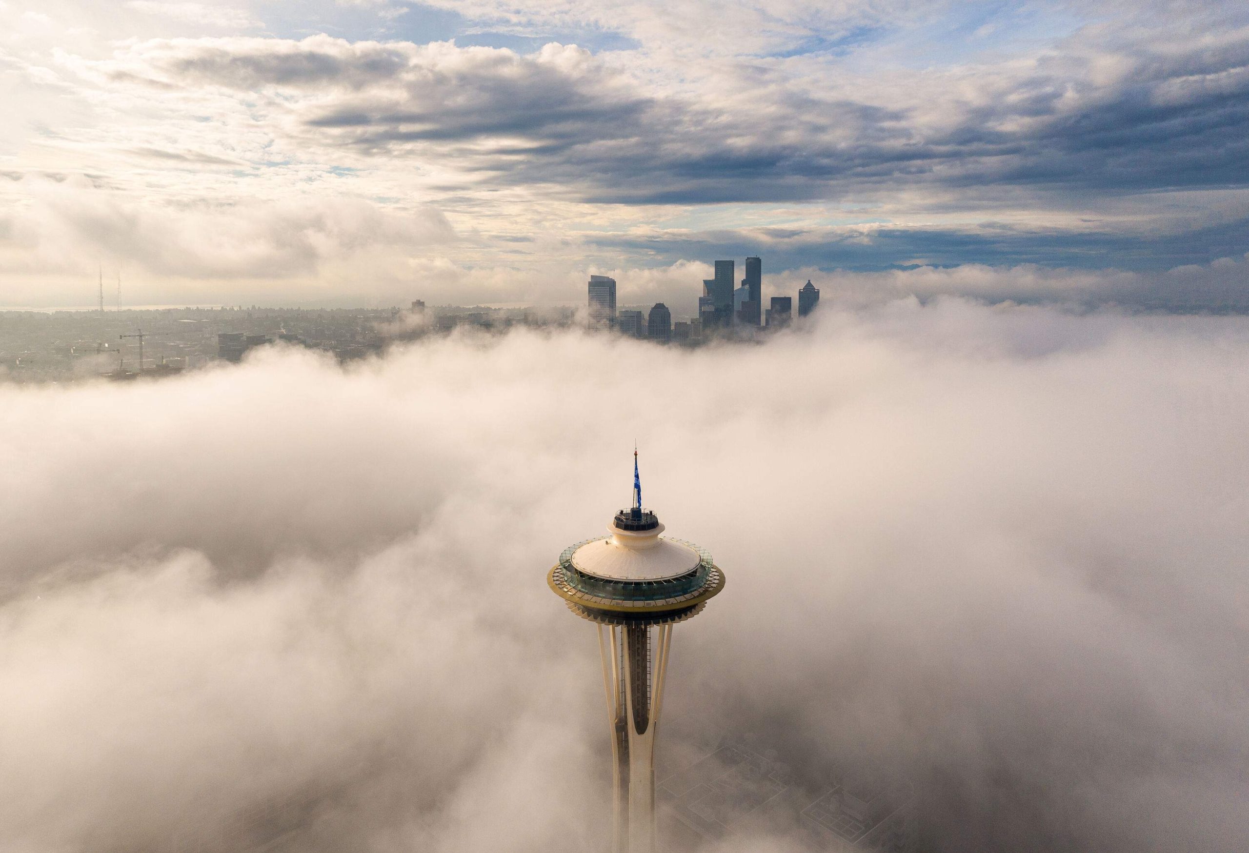 A tall observation tower jutting out on the clouds covering the land with a cluster of tall buildings in the distance.