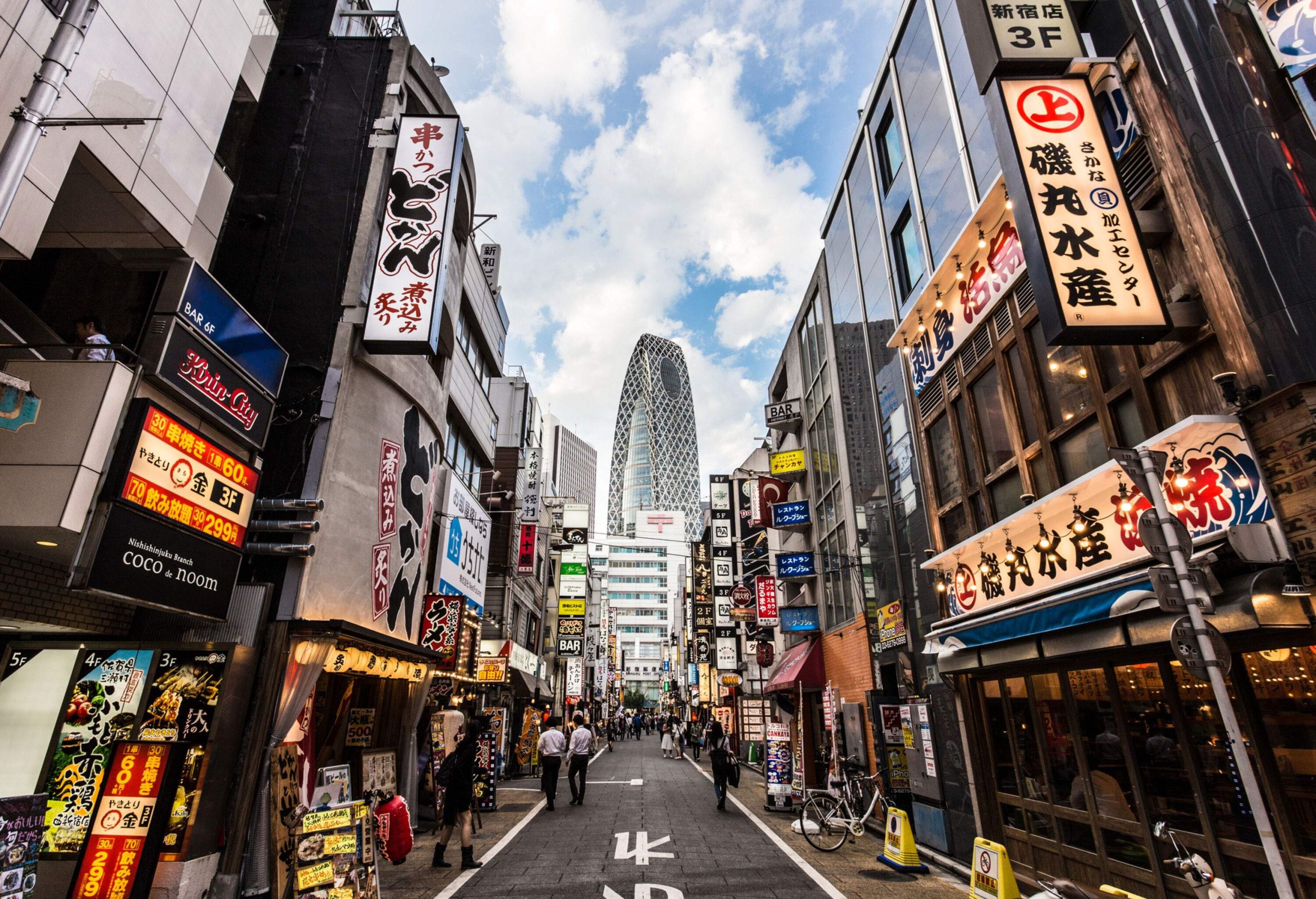 A crowded street lined with commercial establishments with bright LED billboards and views of a tall skyscraper in the background.