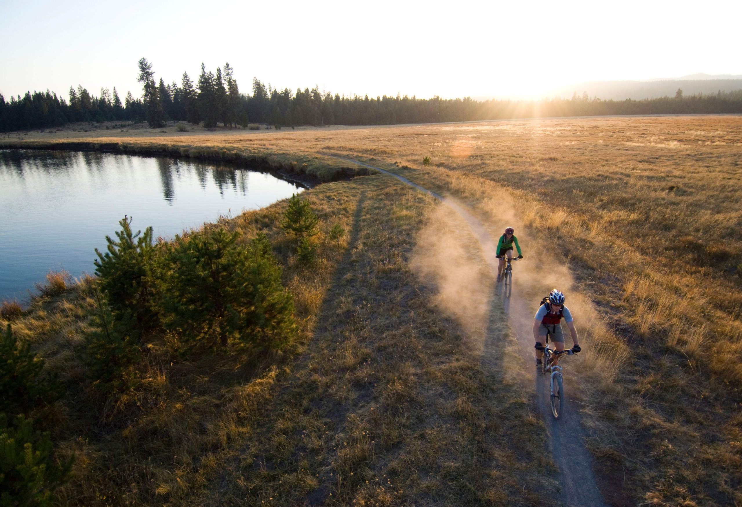 Two cyclists travel on a dusty trail along a calm lake.