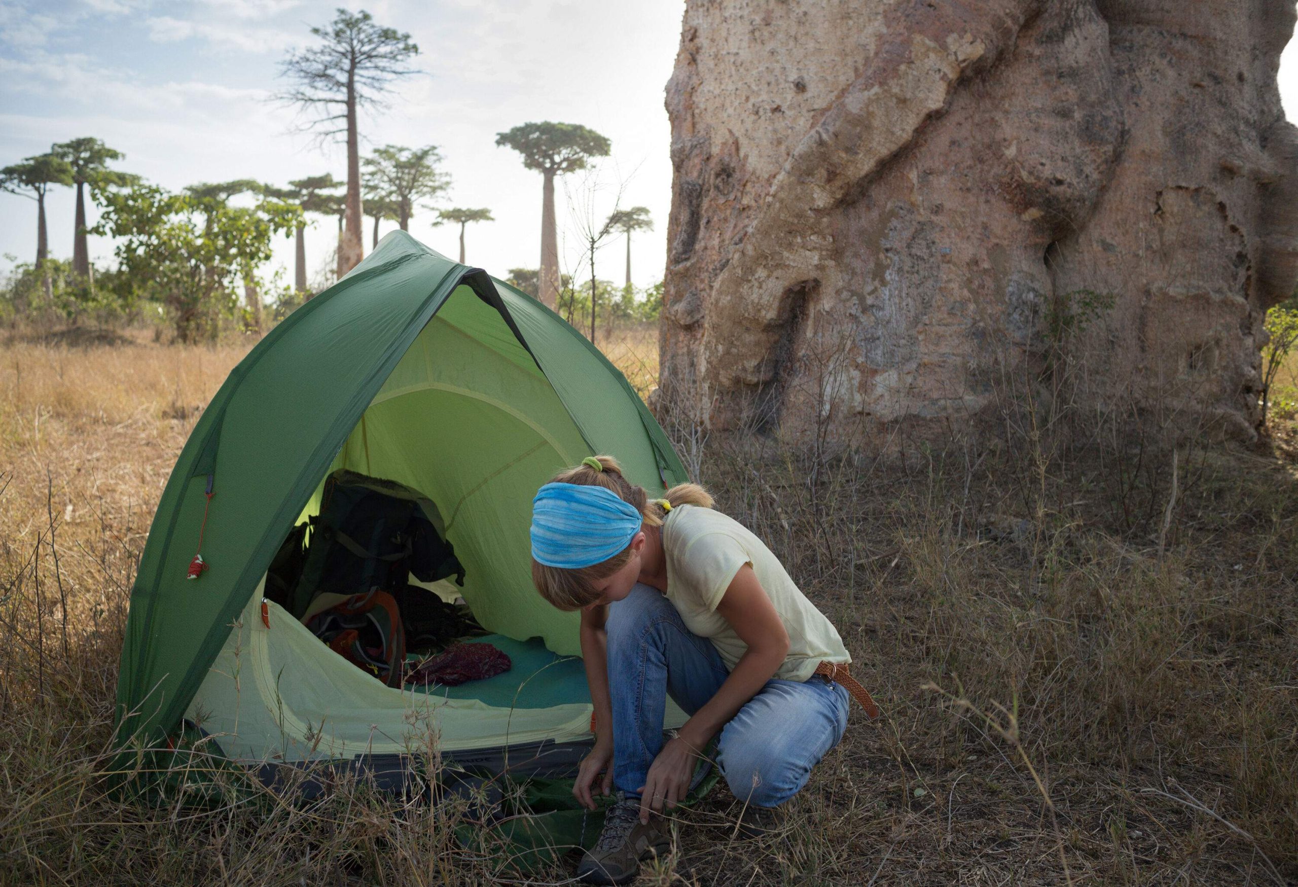 Hiker kneels and ties her shoes beside a tent pitched under a large tree.