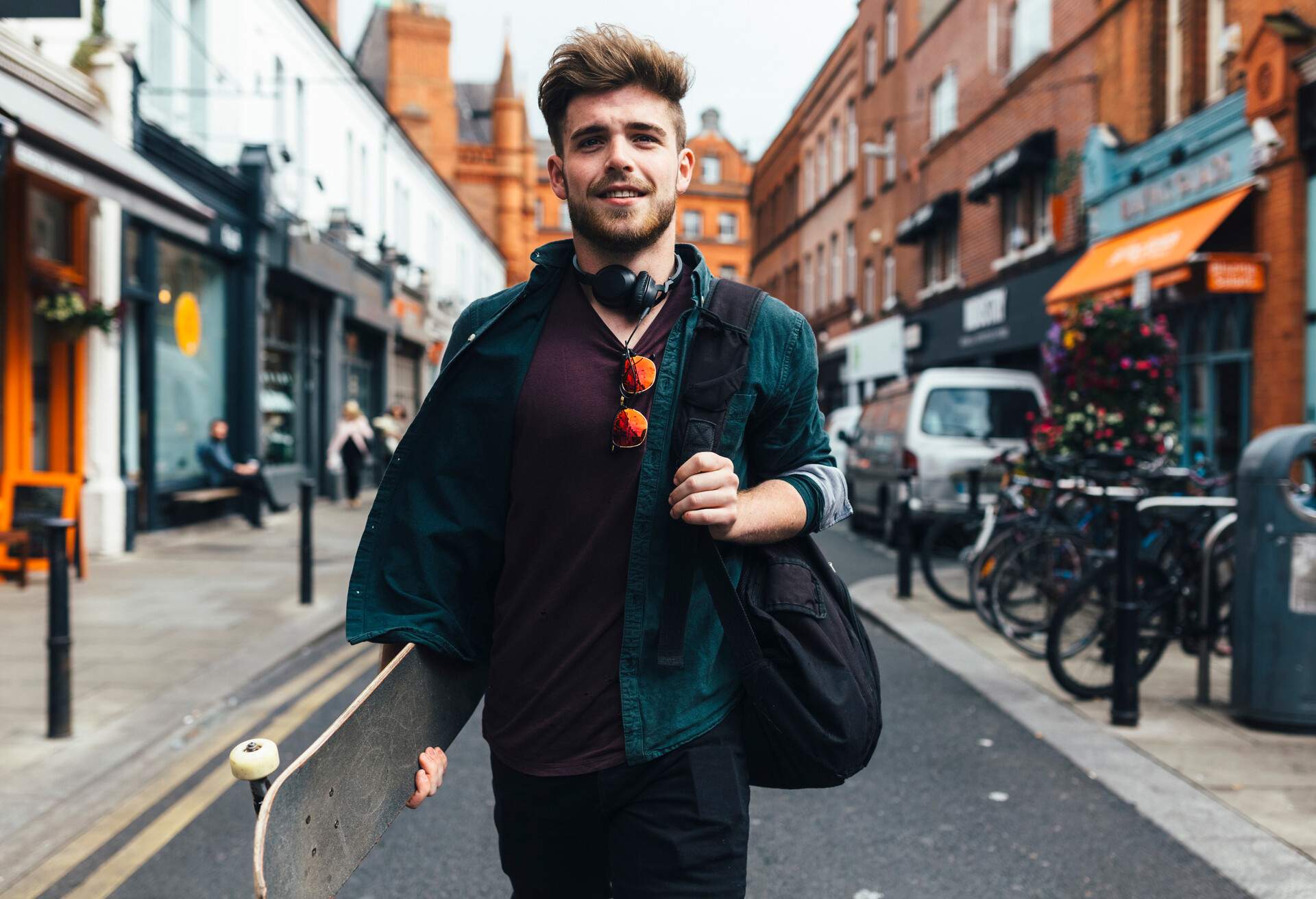 A stylish young man walking down a street, carrying a bag and a skateboard.