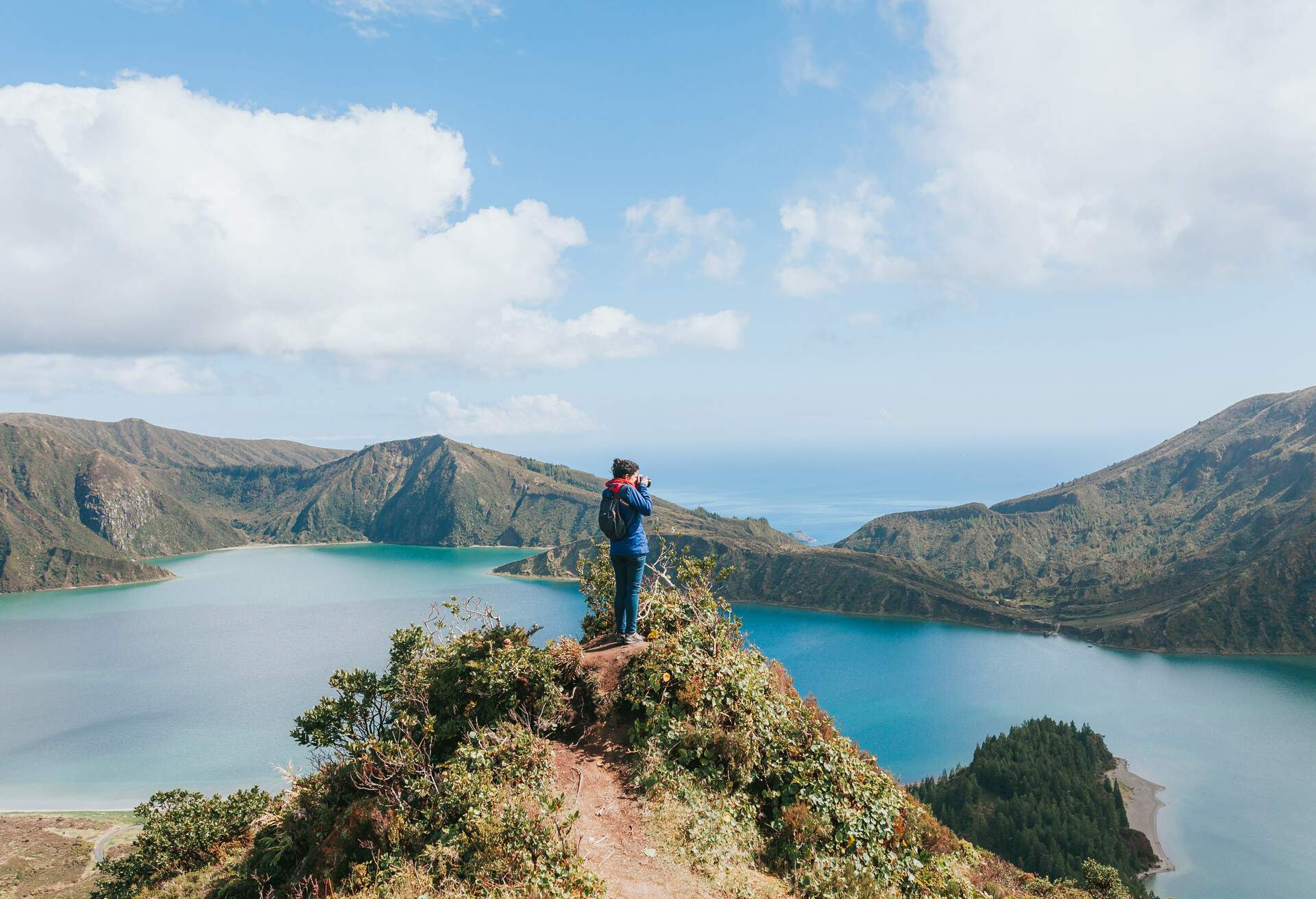 A person stands on a narrow cliff to take a photo of a turquoise lake surrounded by mountains PORTUGAL, AZORES