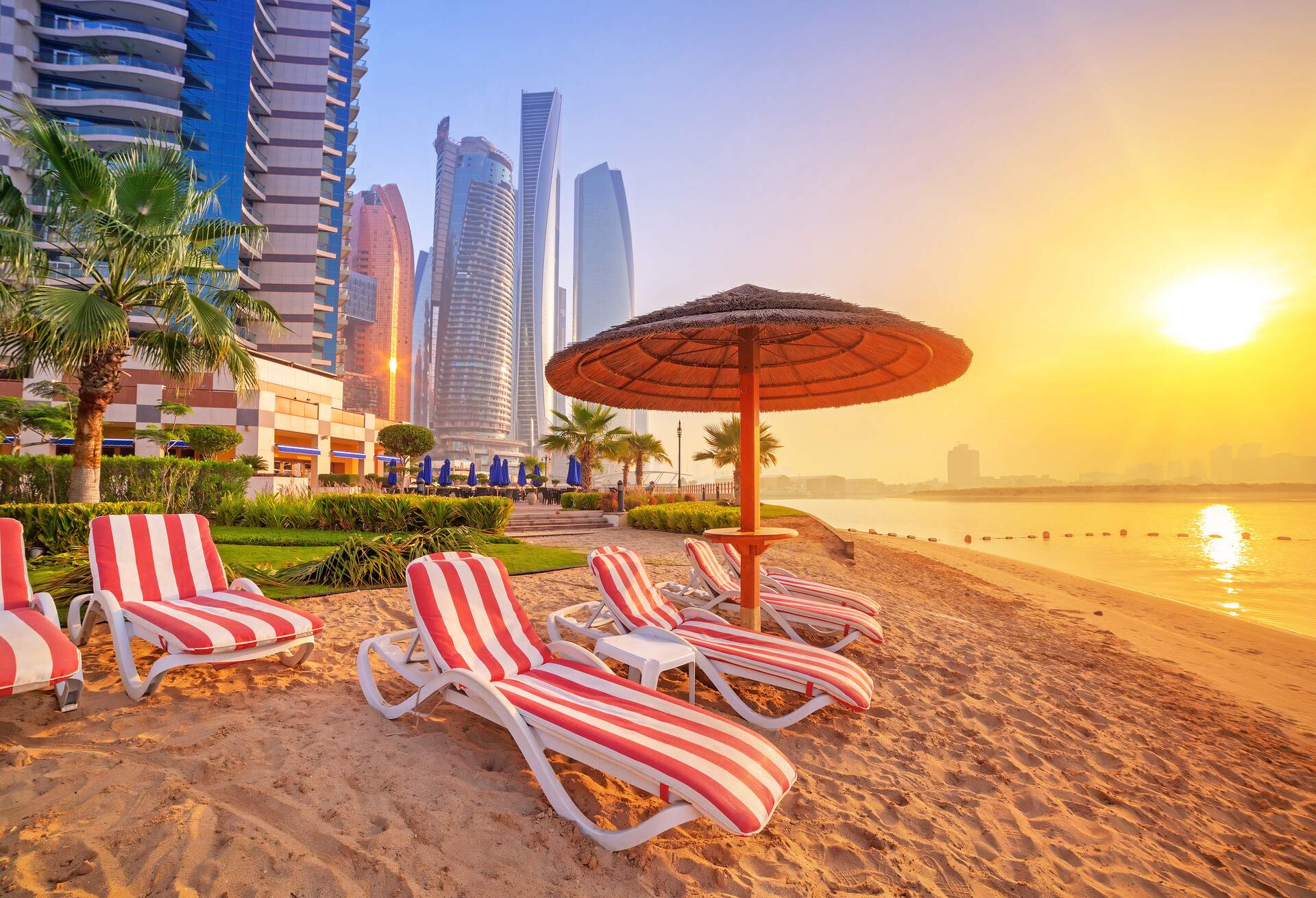 At sunrise, several red-white striped sunbeds under a hut umbrella on a fine sand beach surrounded by tall buildings.