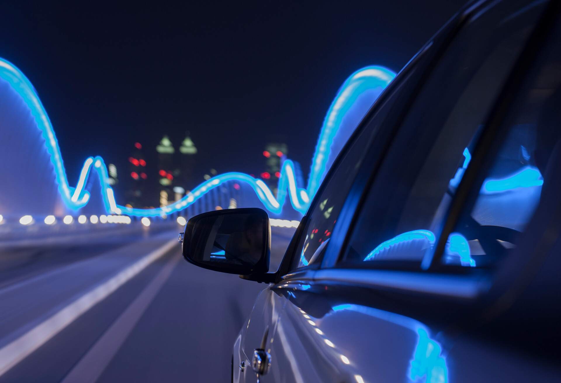 A car driving across a neon-lit bridge at night with lights reflecting on the car's surface.