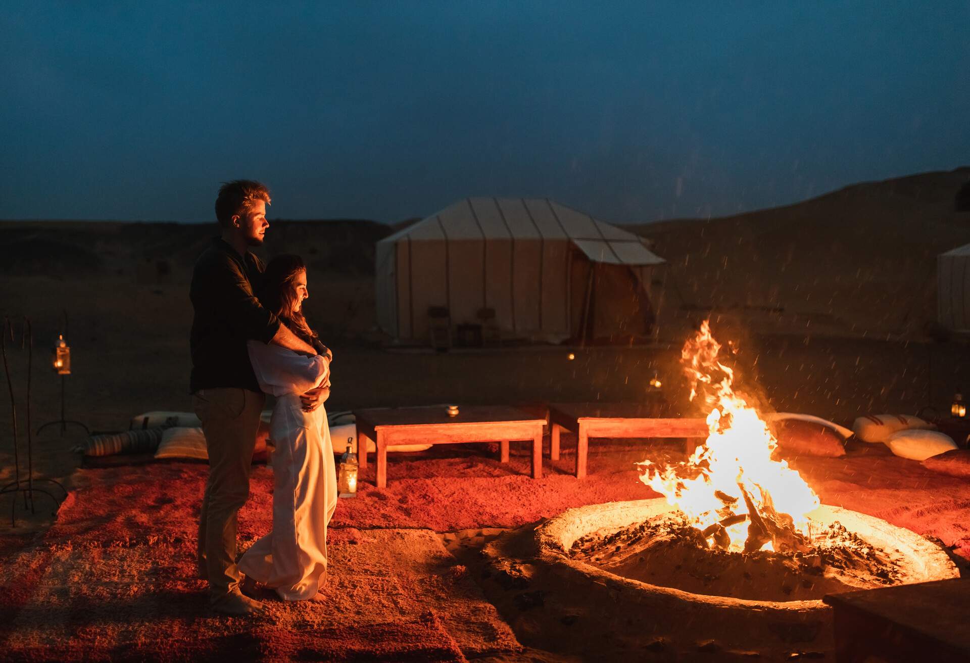 A man hugs a woman from behind enjoying a campfire in the desert.