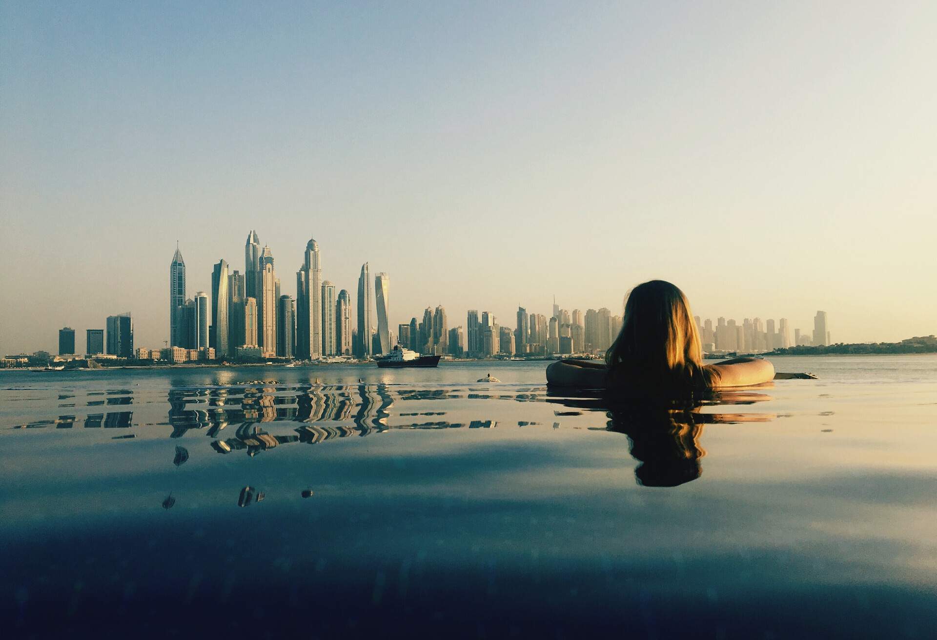 A person immersed in water leans in a pool and gazes at the city skyline.