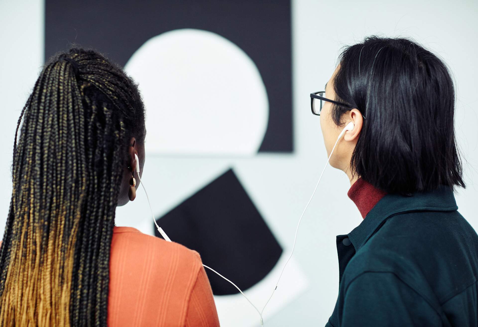 A dark-haired man and a woman with cornrow braids share an earphone while gazing at a piece of wall art.