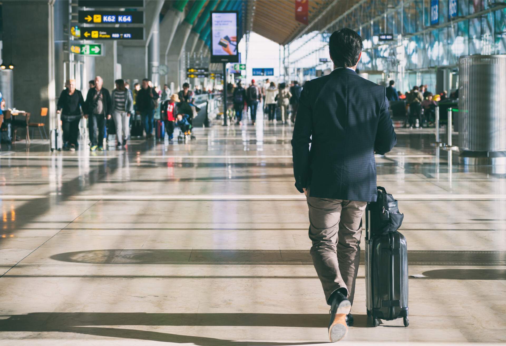 A rear view of a man with a suitcase and many other passengers walking inside the airport corridor.