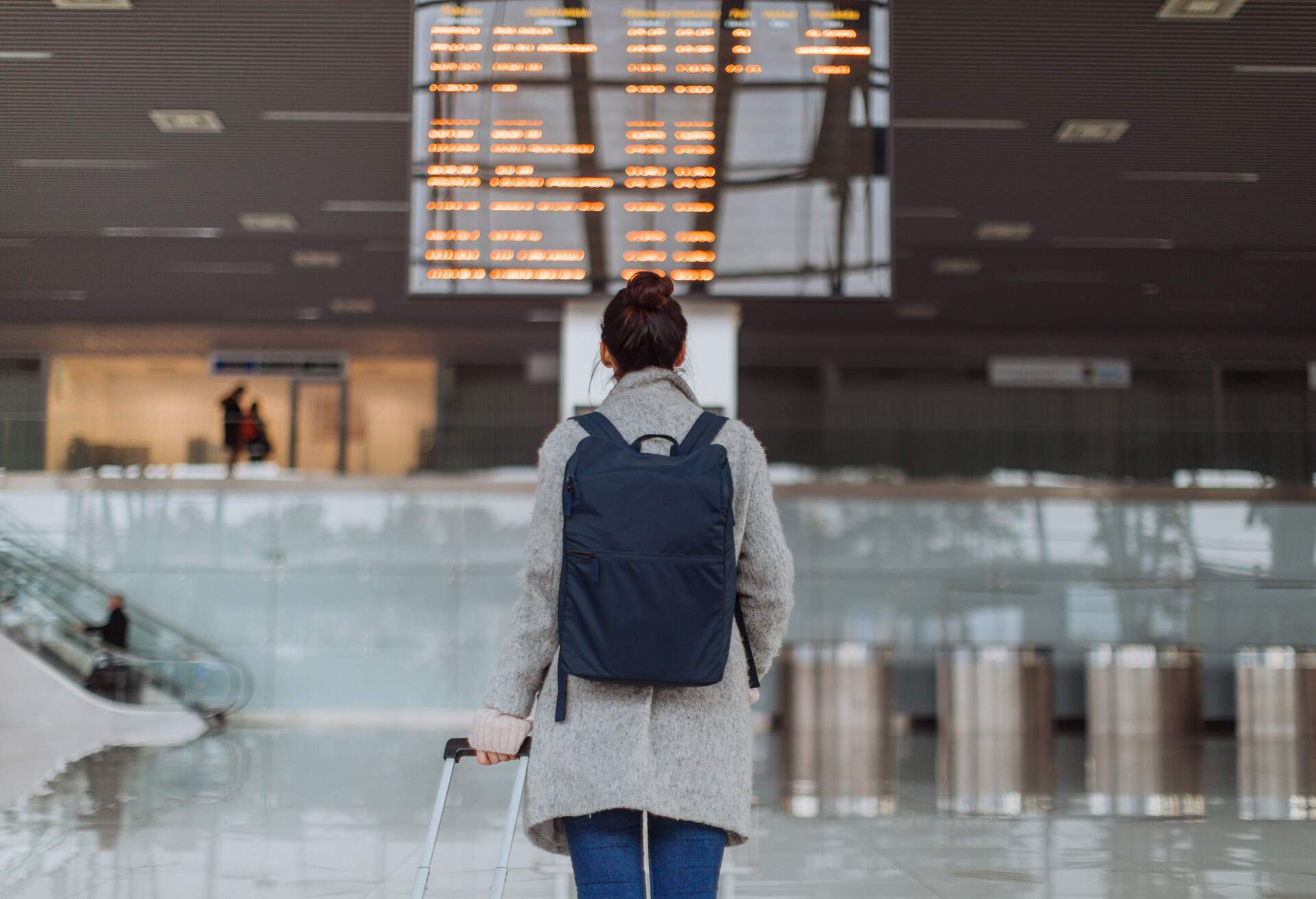 Woman at the airport, checking the arrival and departure boards for flight information while holding her luggage and carrying a backpack.