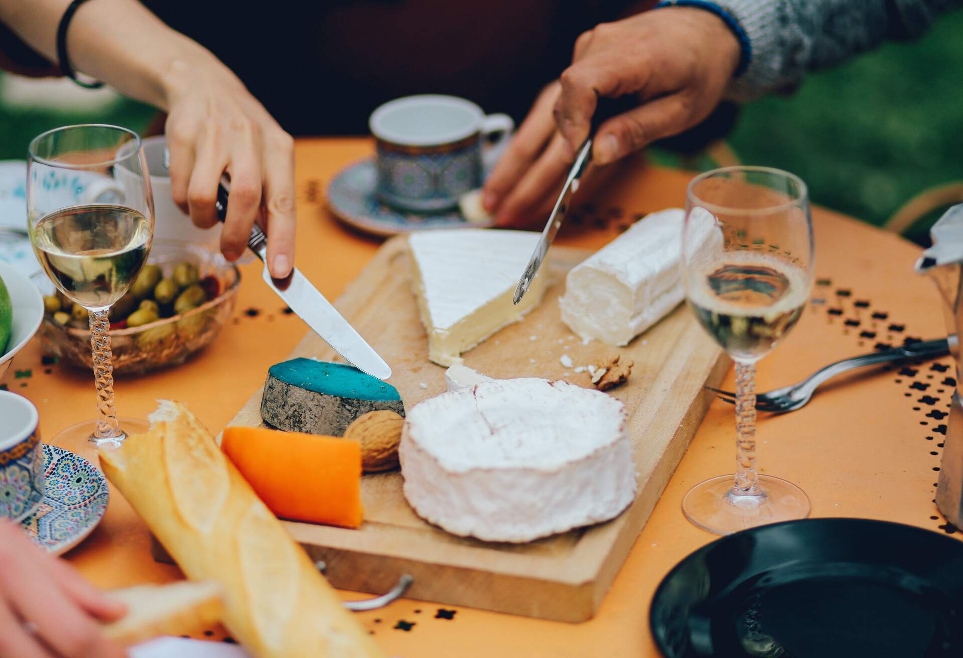 People share a table full of different cheeses, wines, baguette bread, fruits and vegetables.