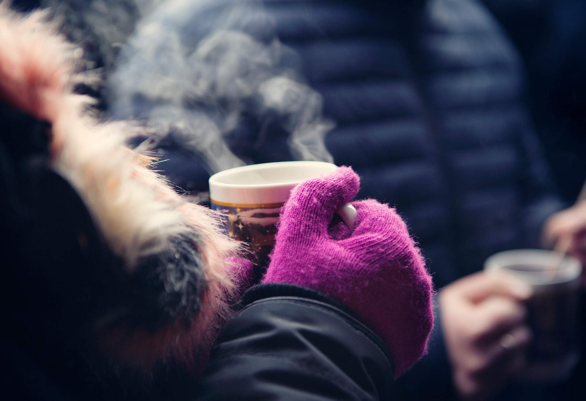 A person wearing winter jacket with purple gloves, holding a steaming mug of hot drink.