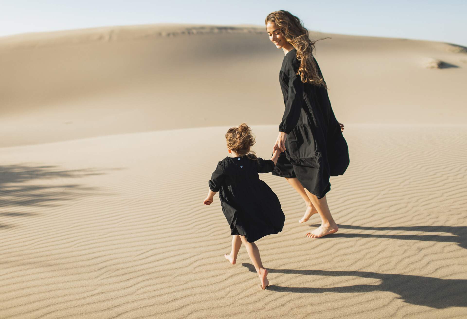 A mother and daughter strolling barefooted on sand dunes in the desert.