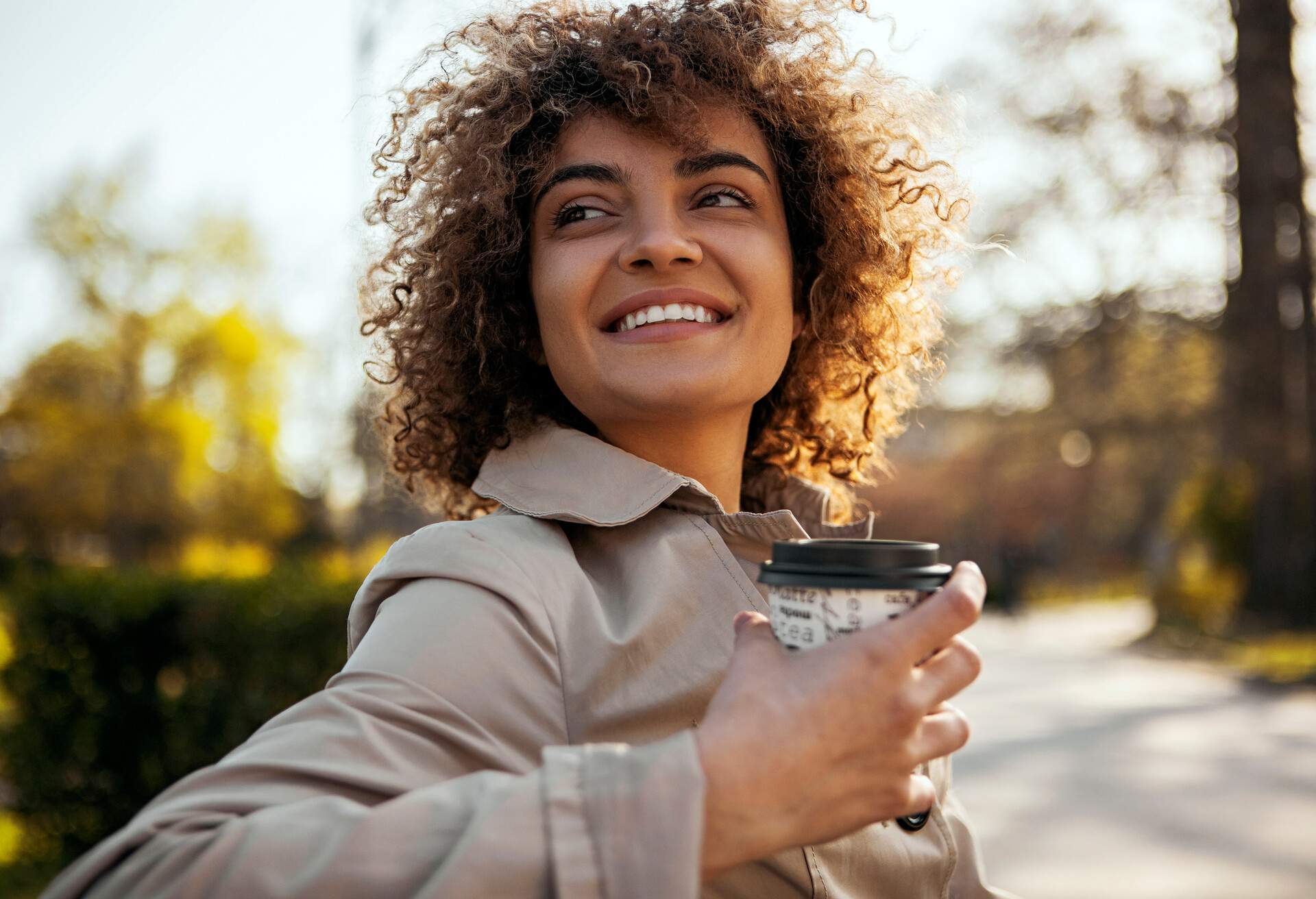 closeup of a smiling woman enjoying her coffee to go on a bench in a park during fall