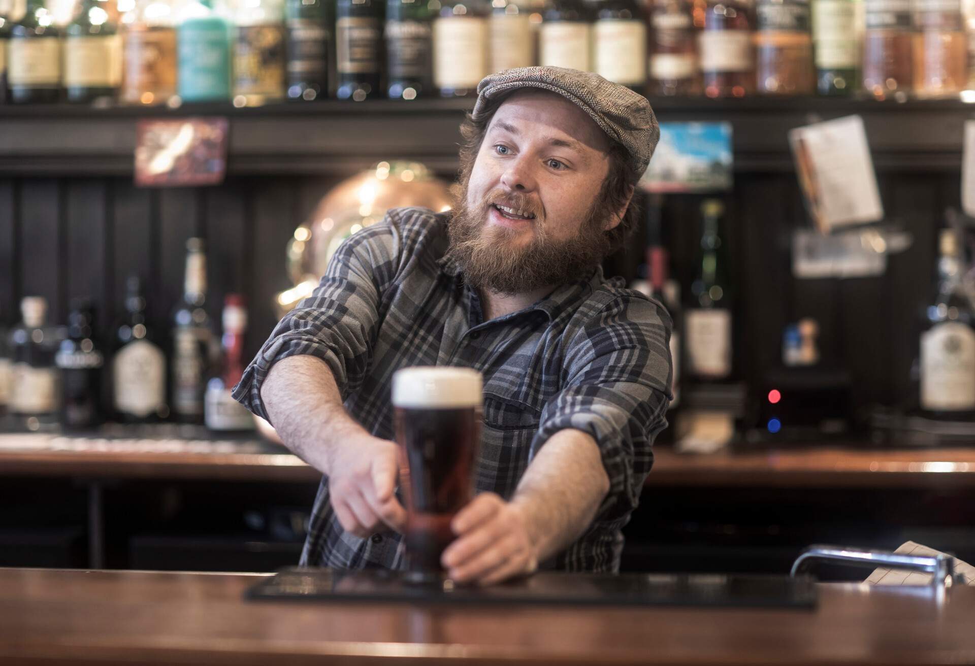 A bartender, with a knowing smile, points to a freshly prepared drink on the counter while scanning the crowd for the awaiting customer.