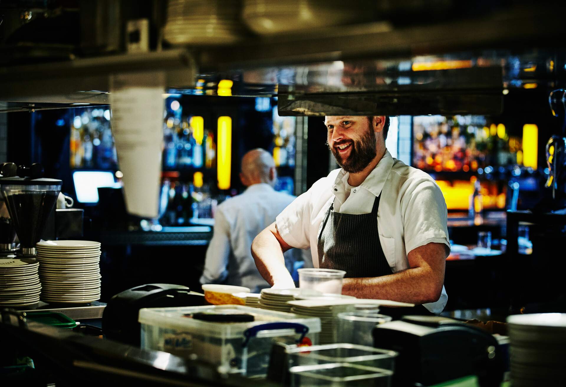 A chef smiling as he works behind a counter lined with stacks of dishes in the kitchen.