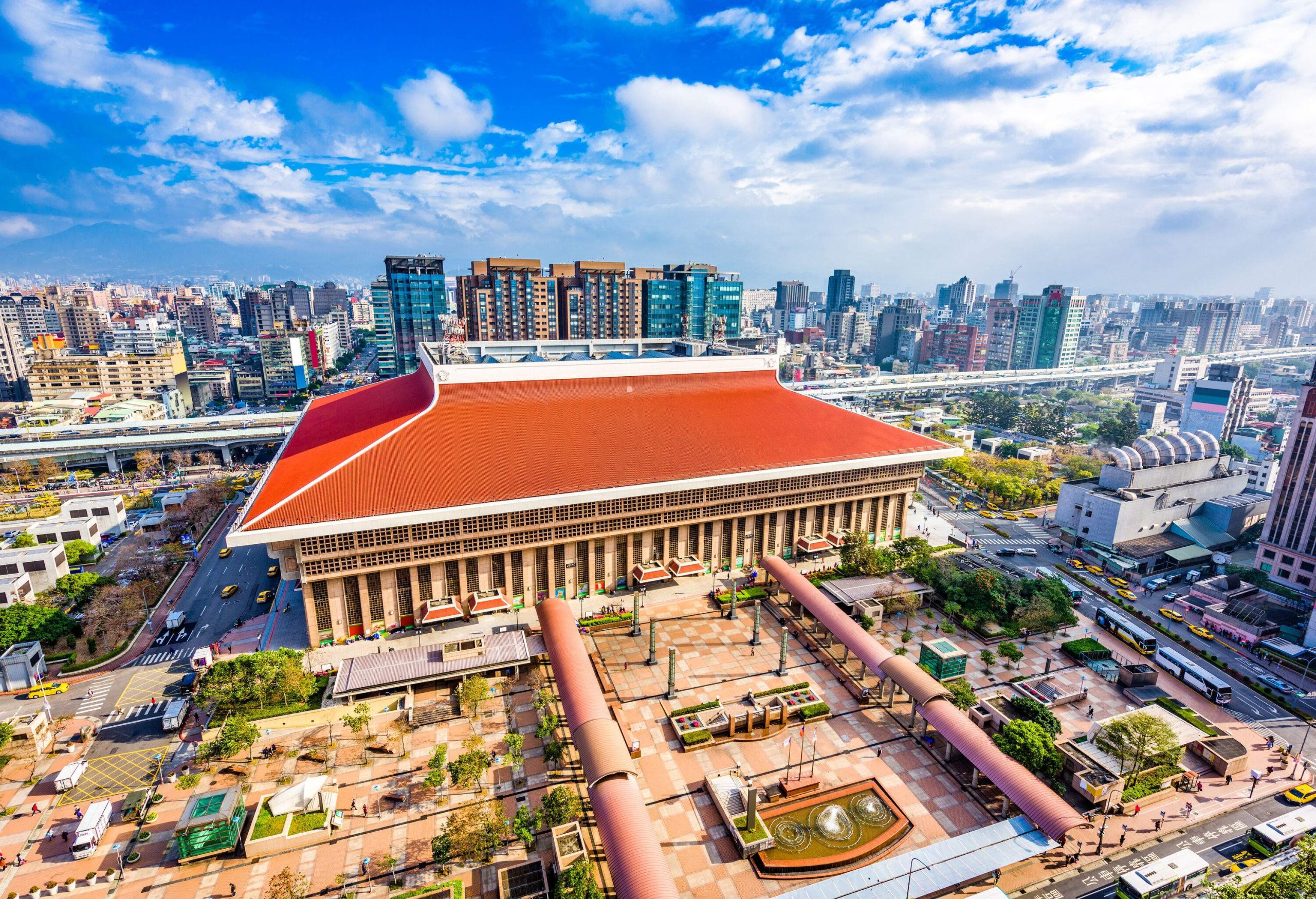 A massive building with red roof and a park with covered walkways against the backdrop of a city skyline.