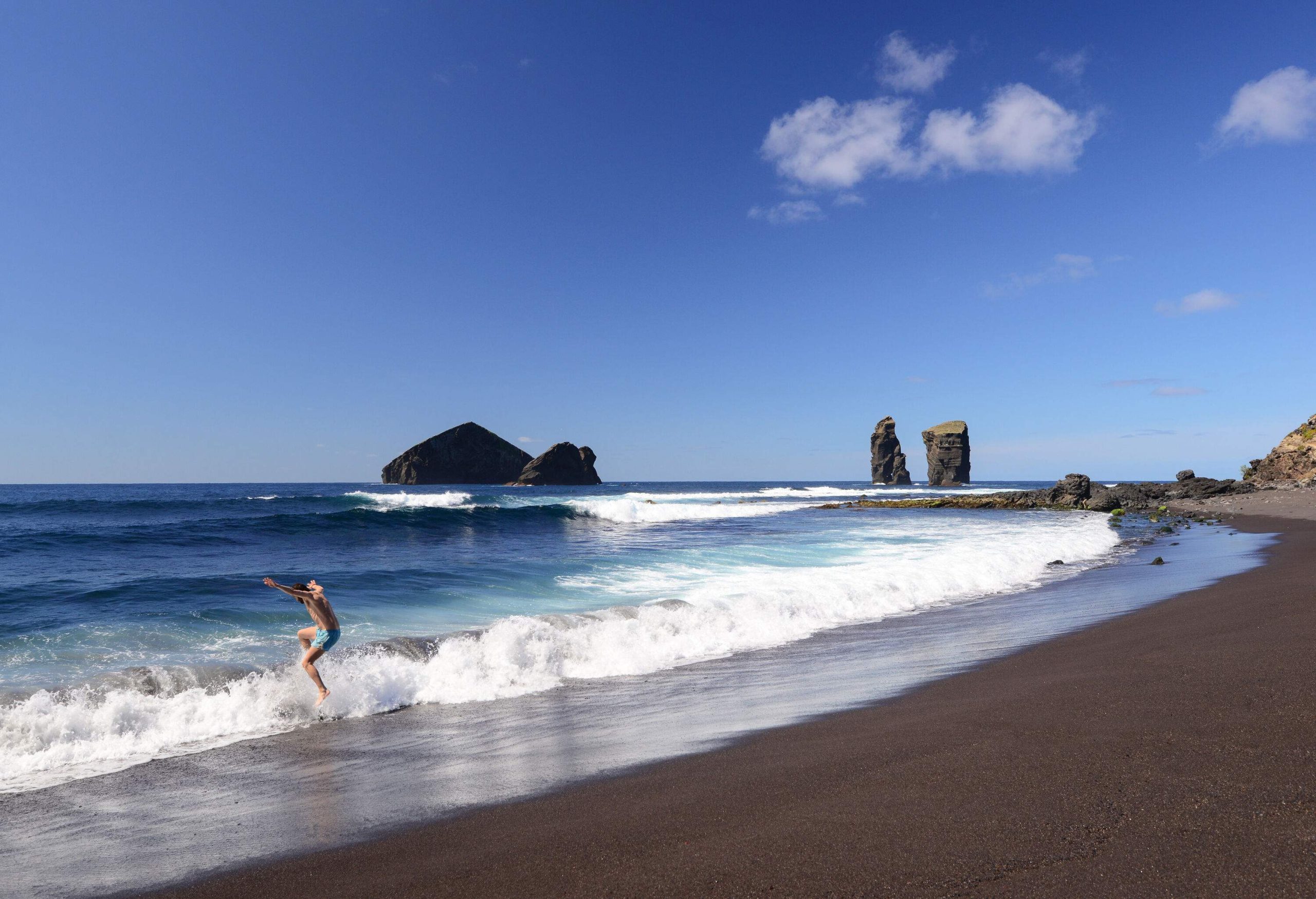 A person jumps over the sea waves in a black sand beach.