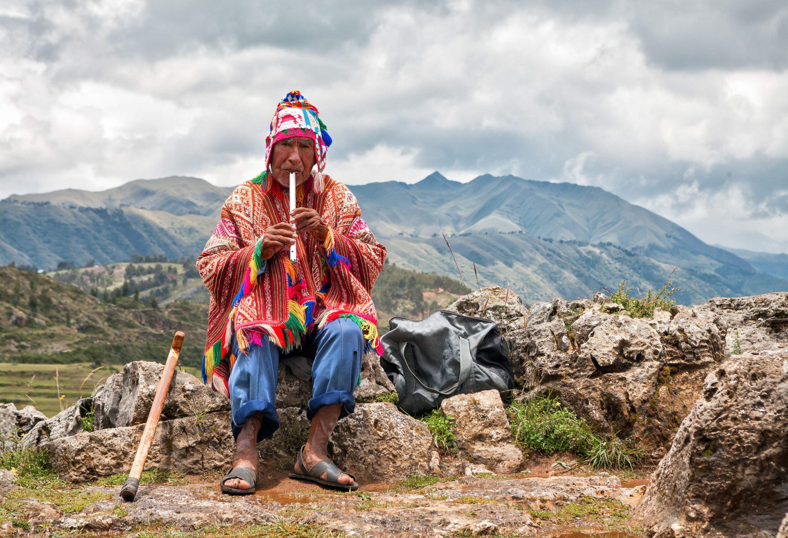 An elderly man draped in a colourful patterned cape playing a musical instrument while sitting on the rocks.