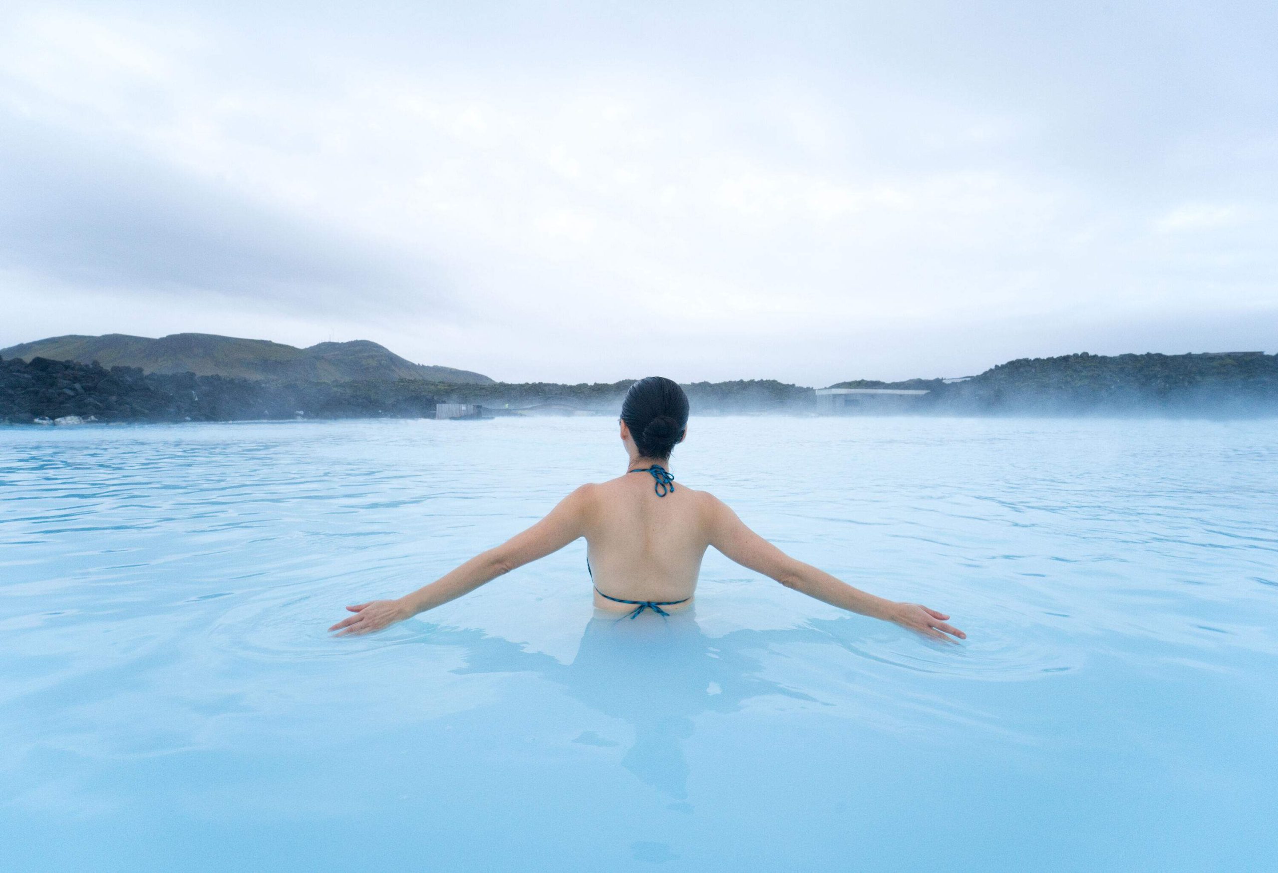 A woman in a bathing suit dips into the milky blue waters of a steaming lagoon.
