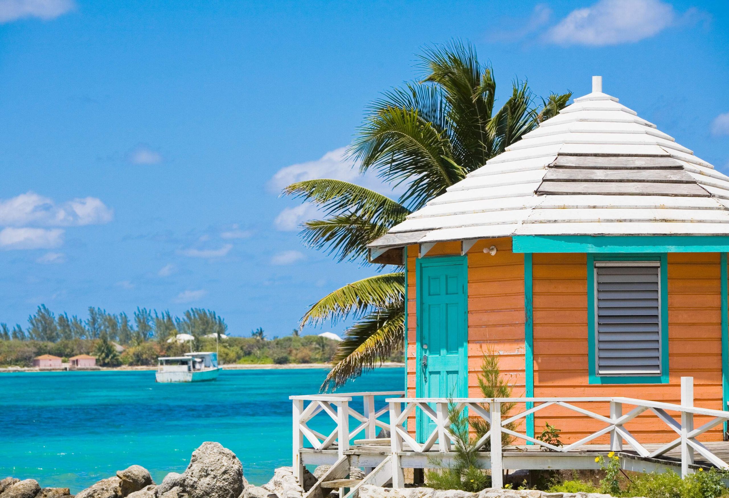 A beach hut on the rocky coast of turquoise waters faces a lone sailing white boat.