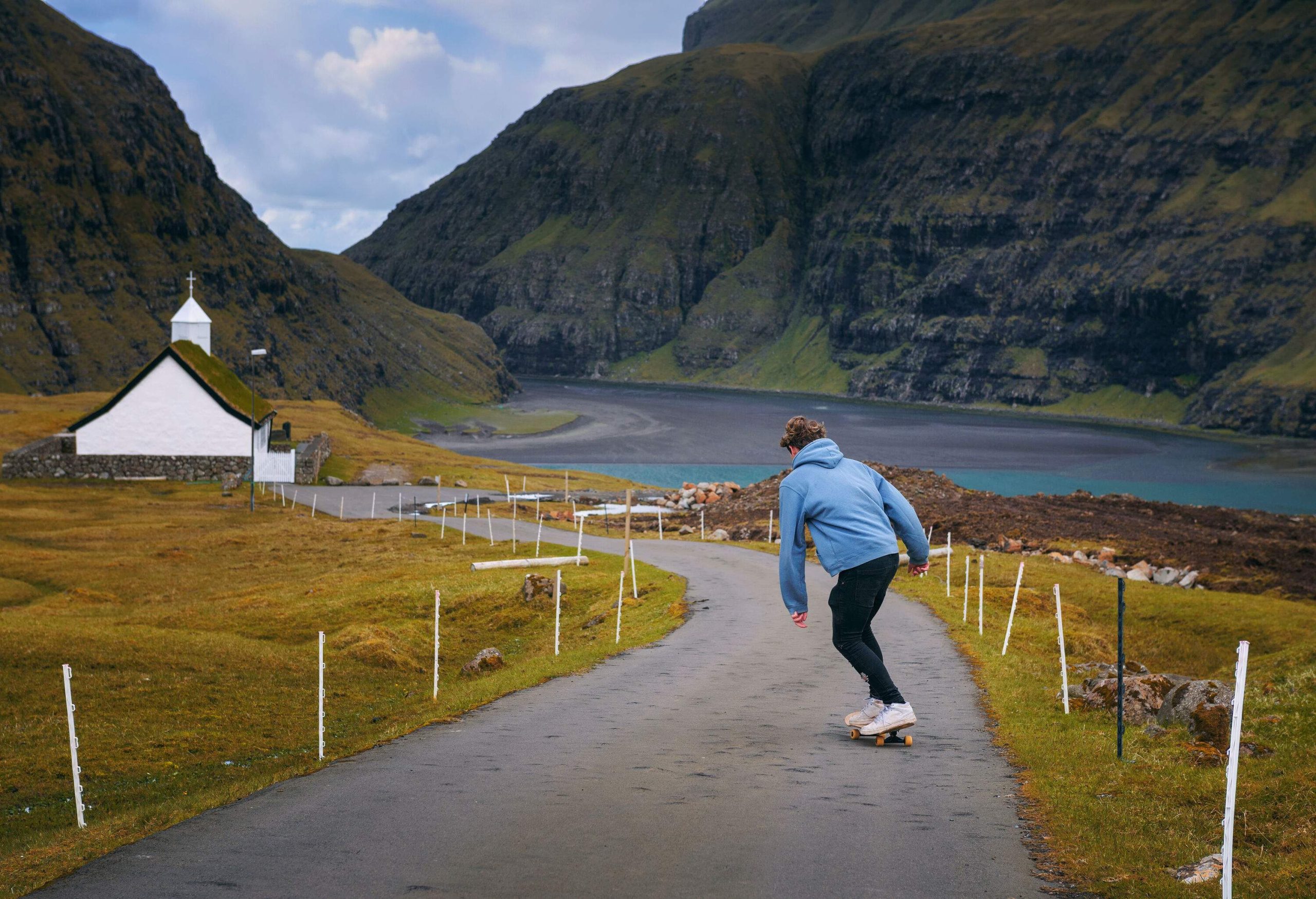 A man in a light blue hoodie rides a skateboard down a winding road towards a white church and calm lake at the base of rugged mountains.