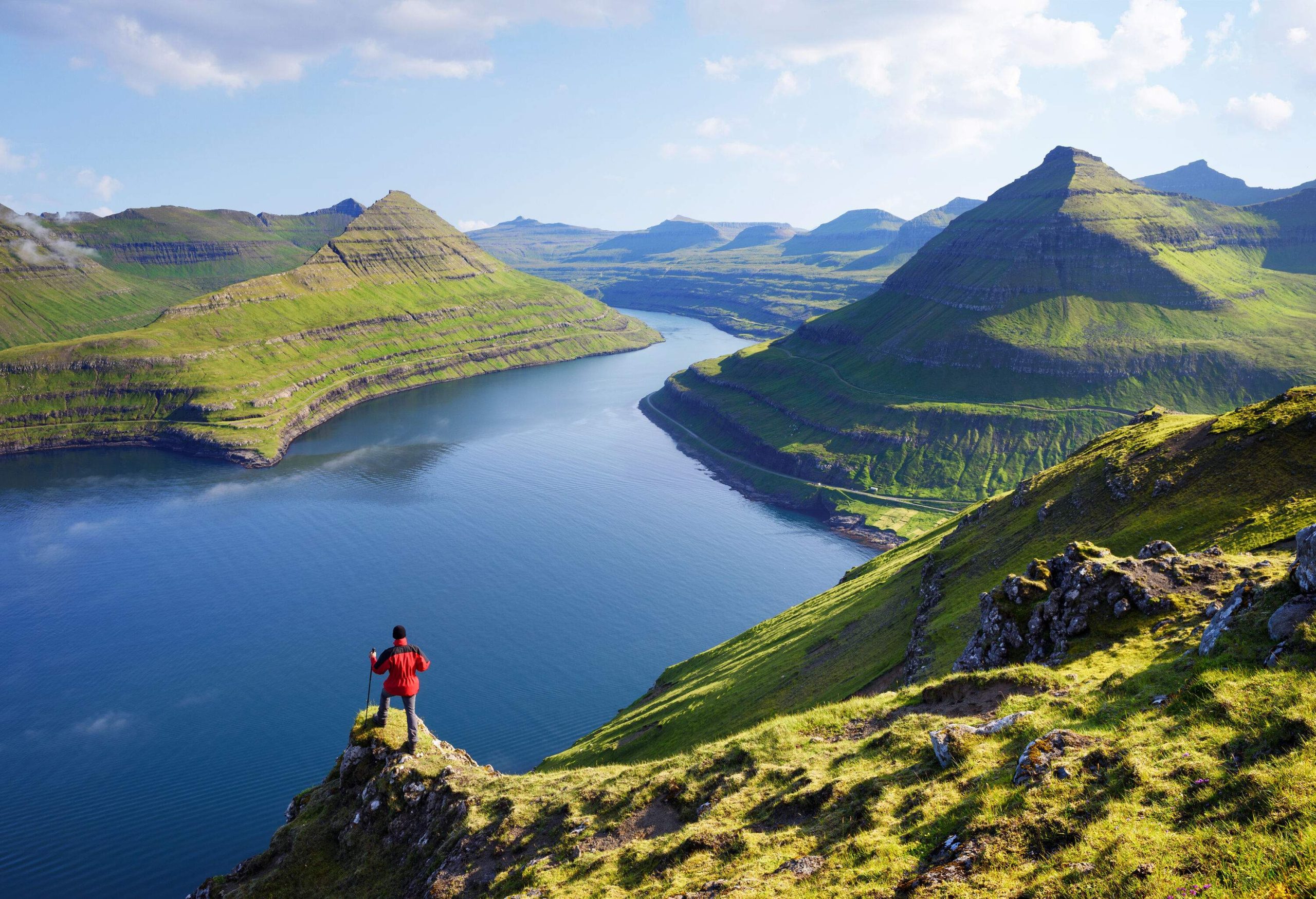 A person in a red jacket standing on a narrow cliff looking at the calm lake beneath the rocky mountains of an island.