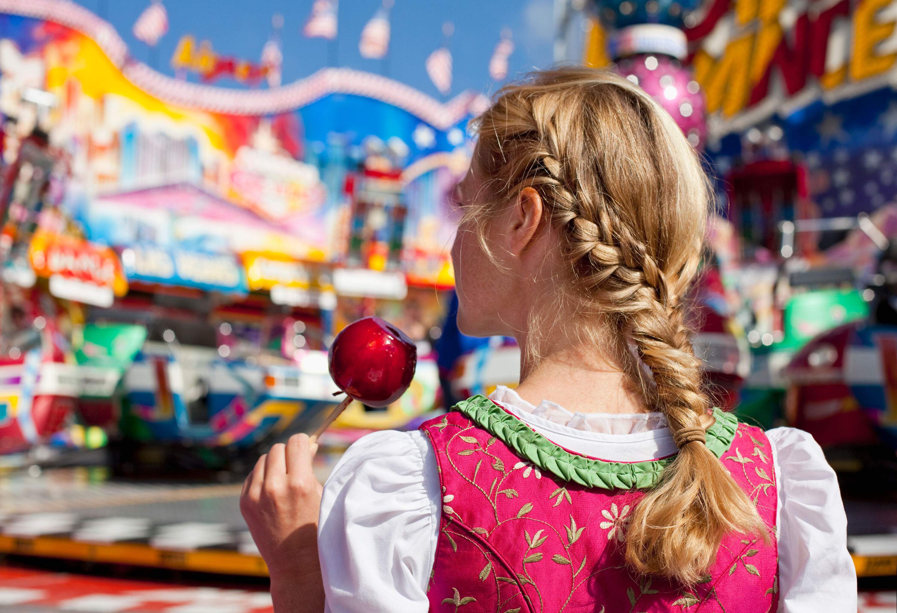 A blonde girl with braided hair holding a sugar-coated apple in a stick.