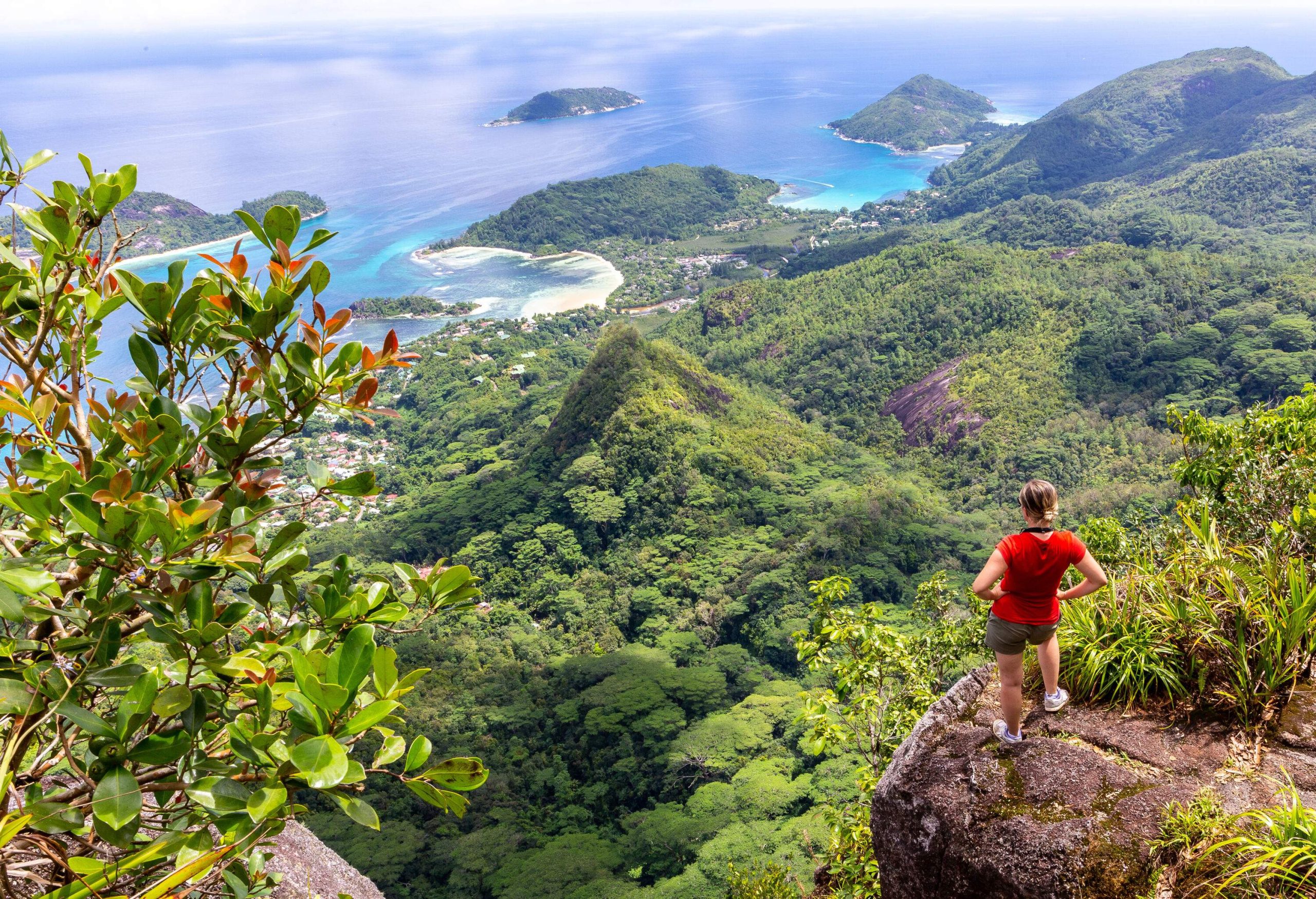 A female hiker in a red shirt standing on a bluff, looking down at the forested hills across the shore.