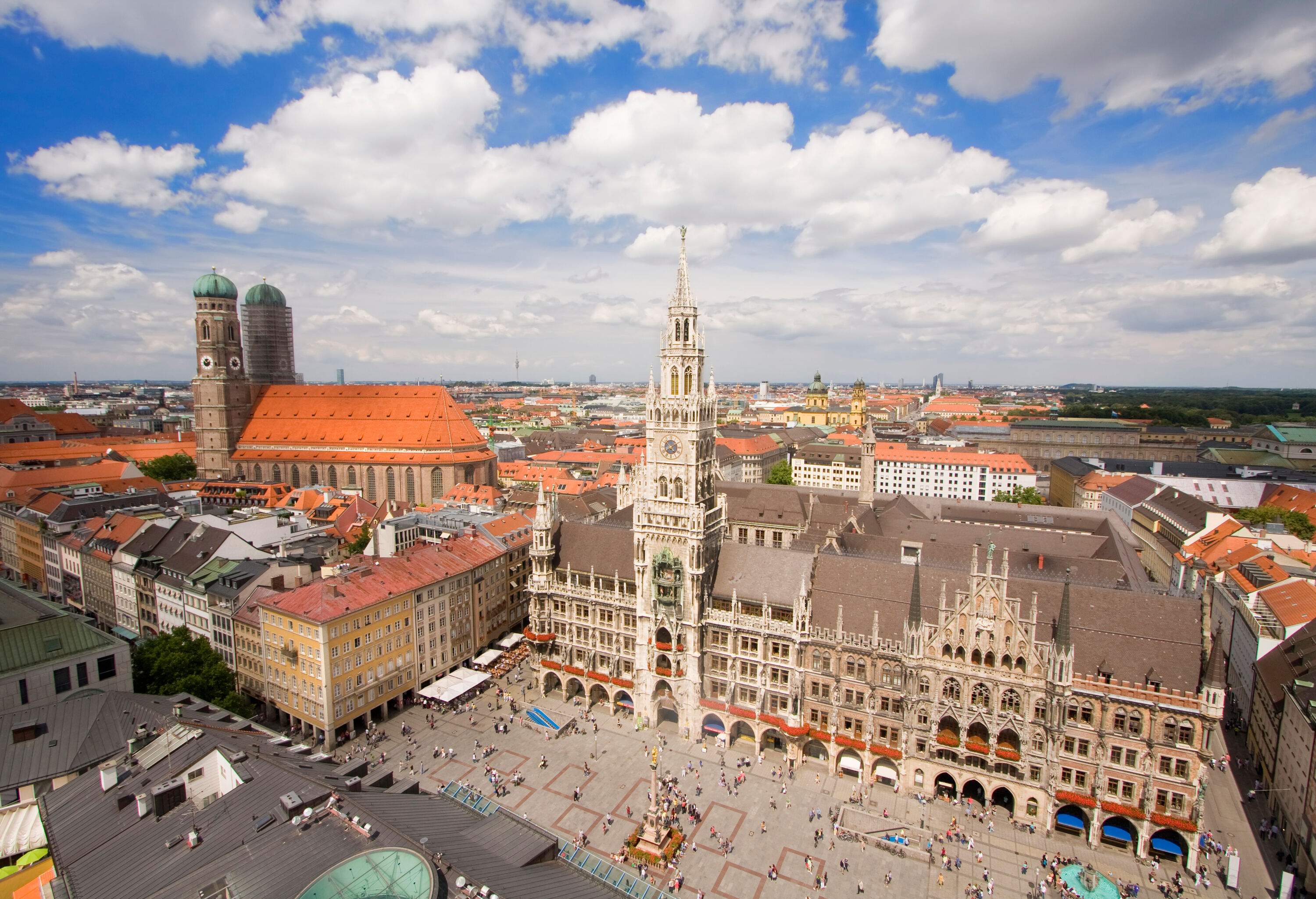 A packed public square in front of a gothic structure with a clock tower and a church with twin towers in the background.