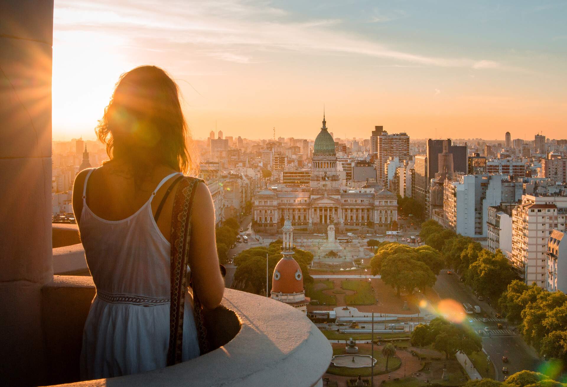 A person stands on a balcony, overlooking the vast cityscape that embraces a serene park below, offering a panoramic view of the dynamic urban landscape.