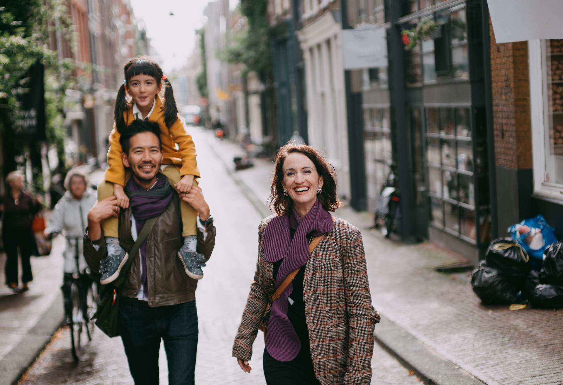 A man with a little girl on his shoulders and a woman smiling as they walk down a wet pavement.