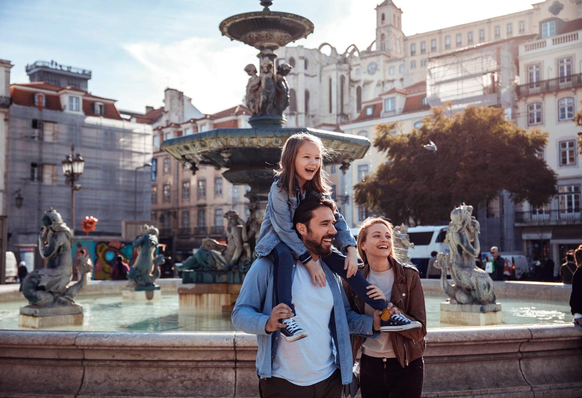 A man carries a kid on his shoulders while standing beside a woman, with a large fountain at the back.