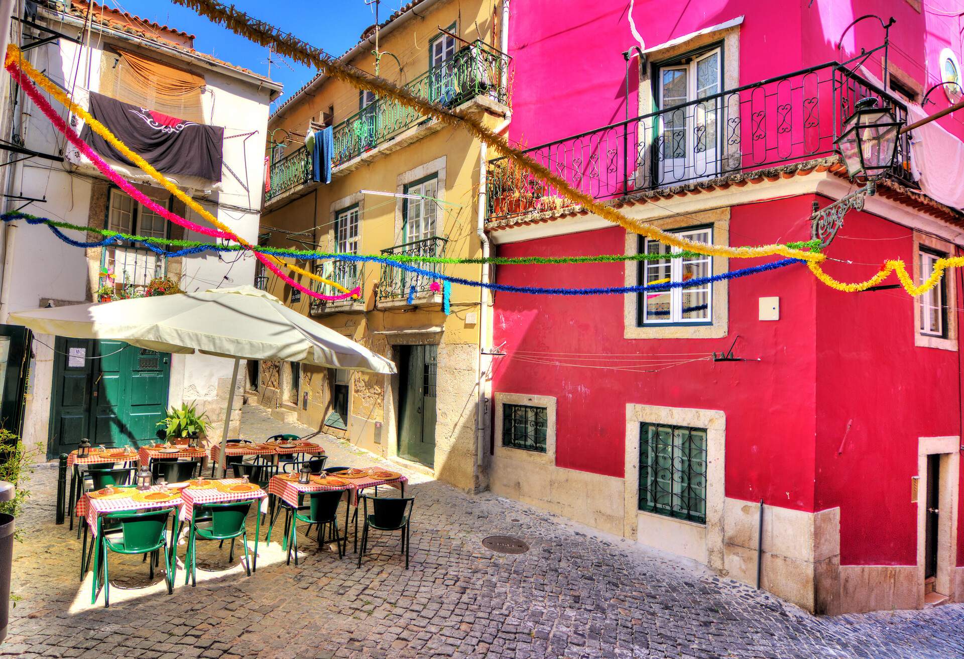 Tables and chairs under an umbrella with dinnerware arranged on a cobbled front yard surrounded by vibrant, colourful houses.