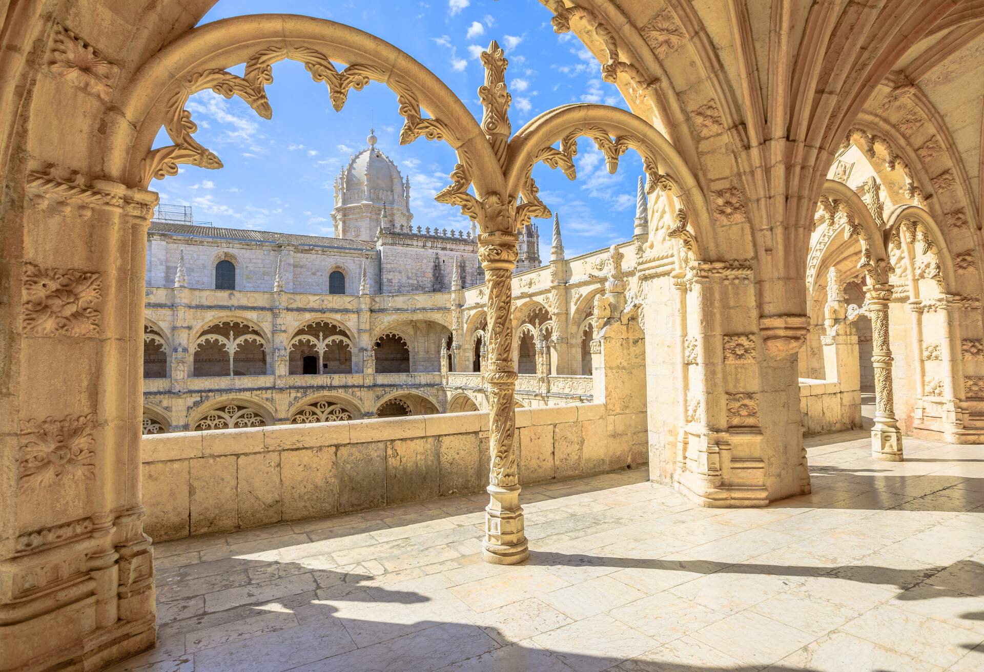 A covered hallway with intricately carved pillars on arched balconies with views of a dome church in the distance.