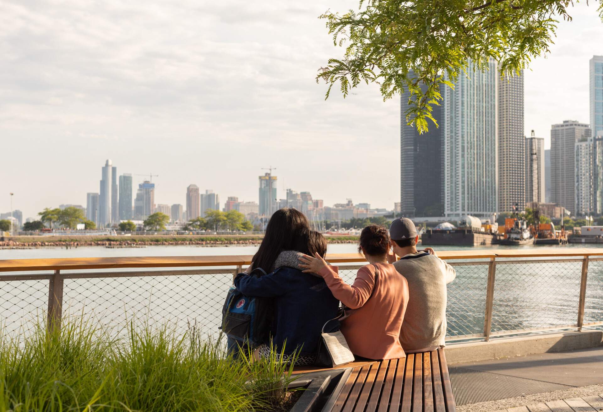 Several people relax in leafy, waterside park with tall skyscrapers in the background