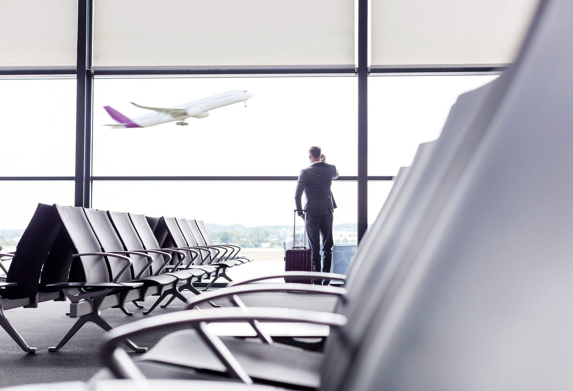 A man in business attire holds his phone in his right ear and stands by the glass walls of an airport departure area.