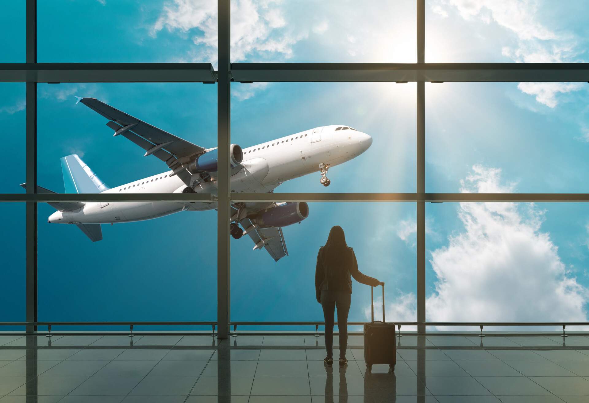 A young woman with a suitcase is standing in the departure hall at the airport, watching an aeroplane take off outside.